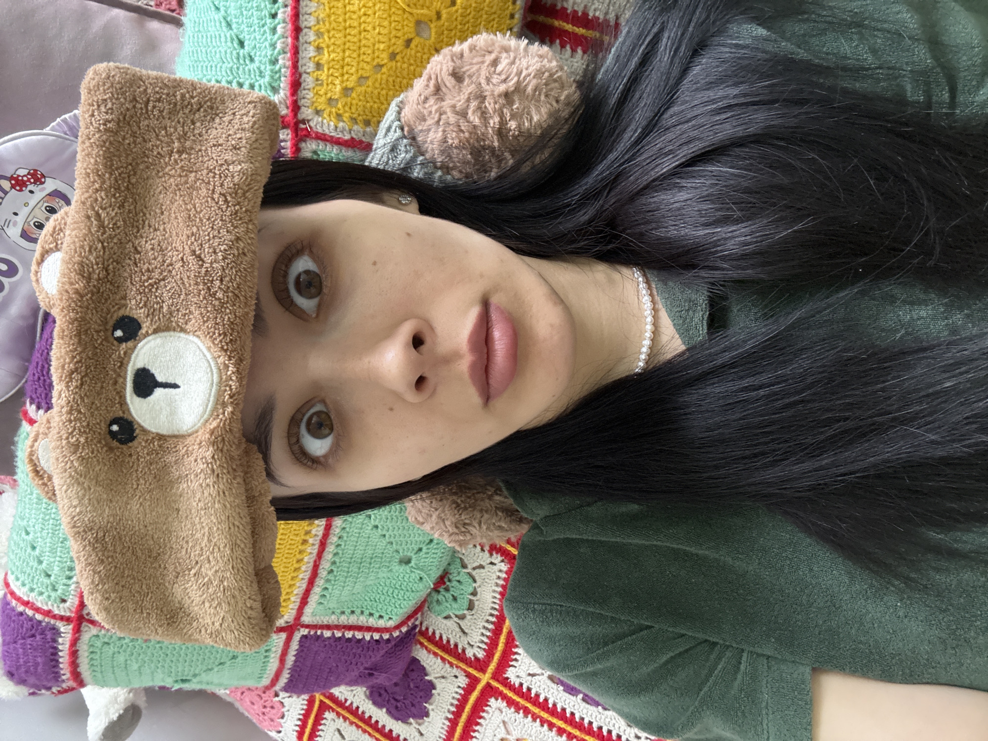 A young woman with long, straight black hair lies on a colorful crocheted blanket. She is wearing a brown bear-shaped headband. A pearl necklace is visible around her neck. She is wearing a green top. The background includes a Hello Kitty bag. There is no information to suggest the picture was taken in Votkinsk, Russia.