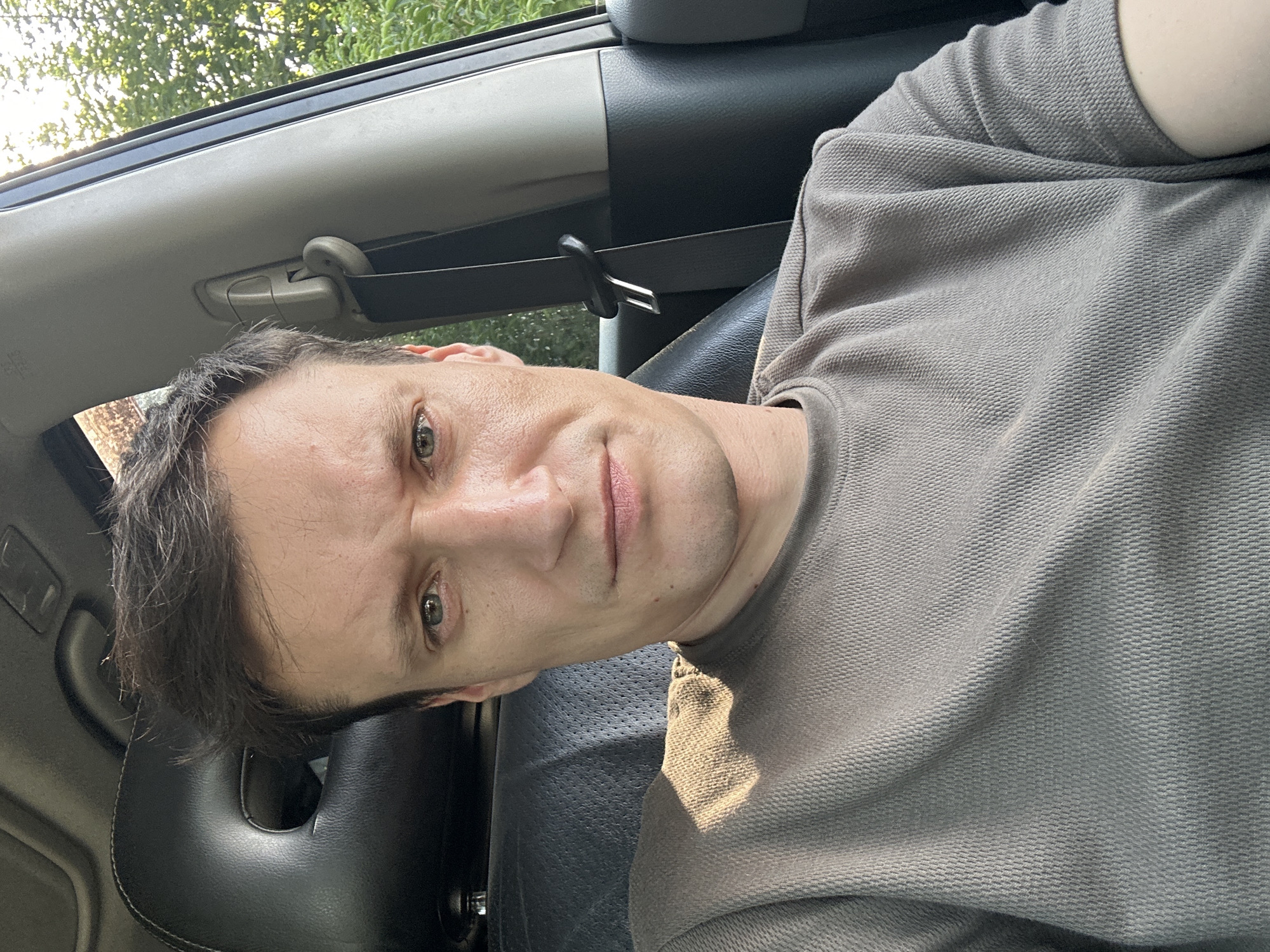 A man is sitting in the driver's seat of a car, facing the camera. He has short, dark brown hair and fair skin. His expression is serious, with his eyebrows furrowed. He is wearing a grey, short-sleeved t-shirt. The interior of the car is visible, including the black leather headrest of the seat behind him and part of the dashboard and door frame. Outside the car window, trees and foliage are visible, suggesting an outdoor, possibly rural setting. There is no indication of the city of Shatava, Ukraine, in the image.
