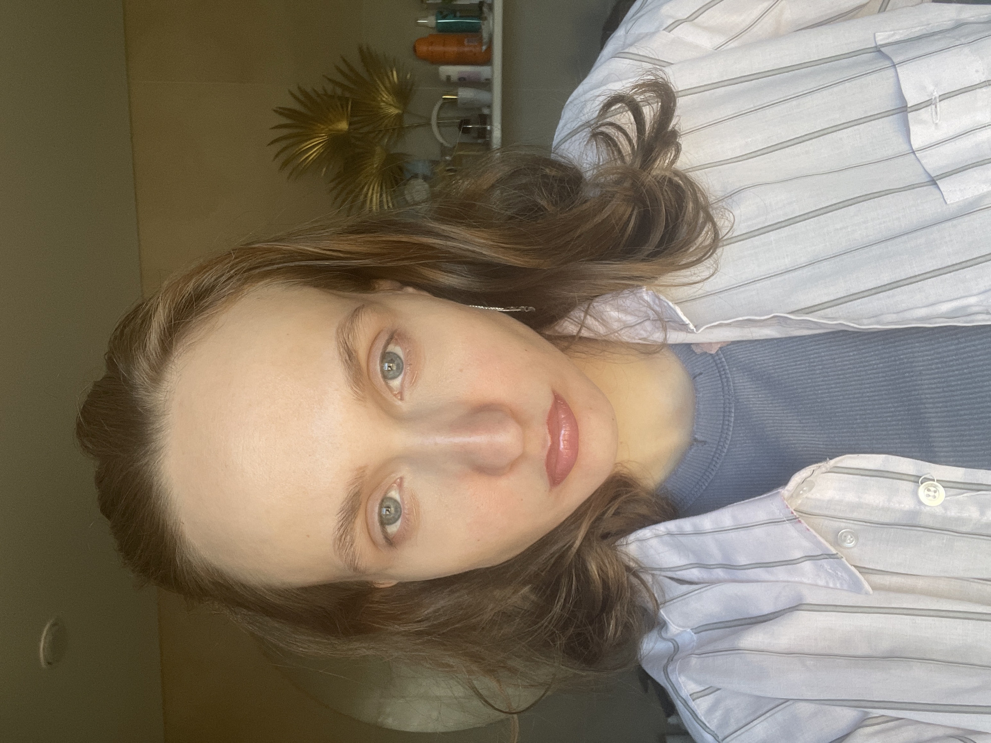A woman with light skin, green eyes, and light brown hair is seen in a close-up, rotated portrait. She has a natural lip color, and a hint of pink blush on her cheeks. She is wearing a blue top under a white and grey striped button-down shirt. Behind her, on a shelf, are several bottles, jars and cans. The background also contains a decorative object resembling a golden sunburst. The location of Podshivalovo, Russia cannot be confirmed from the image alone.