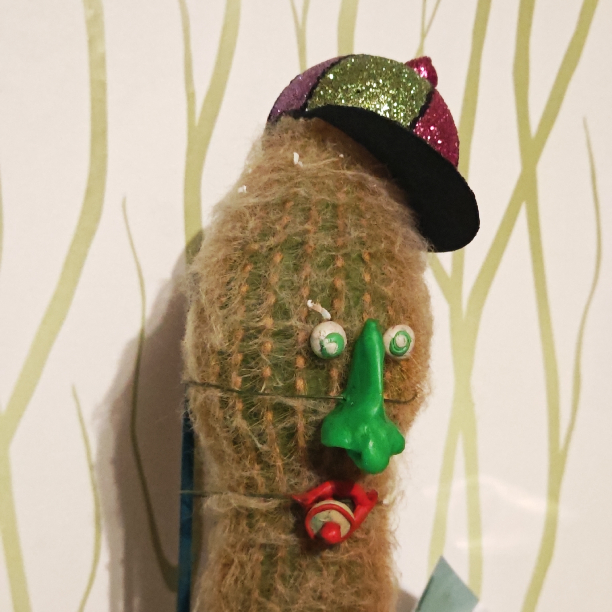 The image displays an anthropomorphic cactus, whimsically decorated to resemble a face. The main subject is a furry, green cactus, possibly a *Mammillaria* species, characterized by its dense, light-brown, hair-like spines.
The cactus is adorned with various objects to form facial features:
- **Headwear:** A small, glittery baseball cap with a black brim and a crown divided into sections of green, pink, and purple glitter is perched atop the cactus.
- **Eyes:** Two small, round, white objects, possibly googly eyes or crafted from clay, with green spiral patterns painted on them, serve as eyes. They are slightly offset from each other.
- **Nose:** A prominent, sculpted green object, likely made of clay or plastic, forms the nose, featuring an irregular, textured shape.
- **Mouth:** A red, sculpted object, possibly clay or plastic, depicts puckered lips with a small red sphere in the center, giving the face a surprised or whimsical expression.
Thin wire or string is visible, wrapped around the cactus, likely securing the facial features in place. A small, blue or teal object is partially visible at the bottom left, possibly part of the cactus's pot or a decorative element.
The setting is indoors, with the cactus positioned against a light-colored wall. The wall features a subtle, repetitive pattern of thin, light green, wavy lines, resembling stylized branches or organic forms. A faint shadow of the cactus is cast on the wall to its left. The lighting appears bright and even, suggesting daytime or well-lit indoor conditions. No specific location cues are visible beyond the general indoor setting. The overall mood conveyed is playful and creative. There is no visible text in the image. The location context is Kladovitsy, Russia.