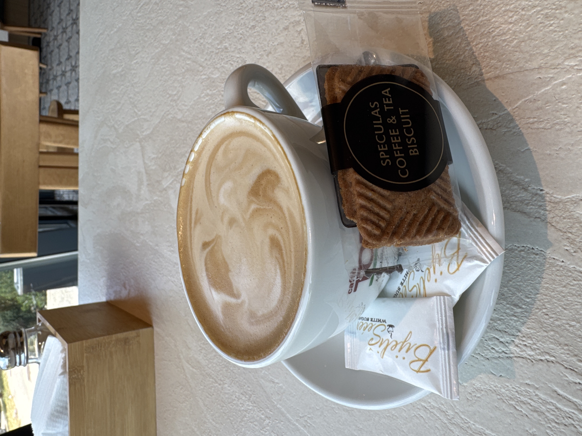 A close-up, eye-level shot captures a white ceramic mug filled with a latte or cappuccino, showcasing delicate latte art on its foamy surface. The mug rests on a matching white saucer, which also holds a packaged "SPECULAS COFFEE & TEA BISCUIT" and two sachets of "Bijelić šećer WHITE SUGAR." The biscuit, brown and intricately patterned, is sealed in clear plastic with a prominent black circular label. The sugar packets are white with gold branding.
The items are placed on a light-colored, textured table surface, illuminated by bright natural light, indicating it is daytime. In the blurred background to the left, wooden furniture or shelving is visible, suggesting an indoor setting. Beyond these interior elements, a hint of an outdoor view with some greenery can be seen through a window. The scene suggests a tranquil coffee break in Bjeliši, Montenegro. No people are visible in the image.