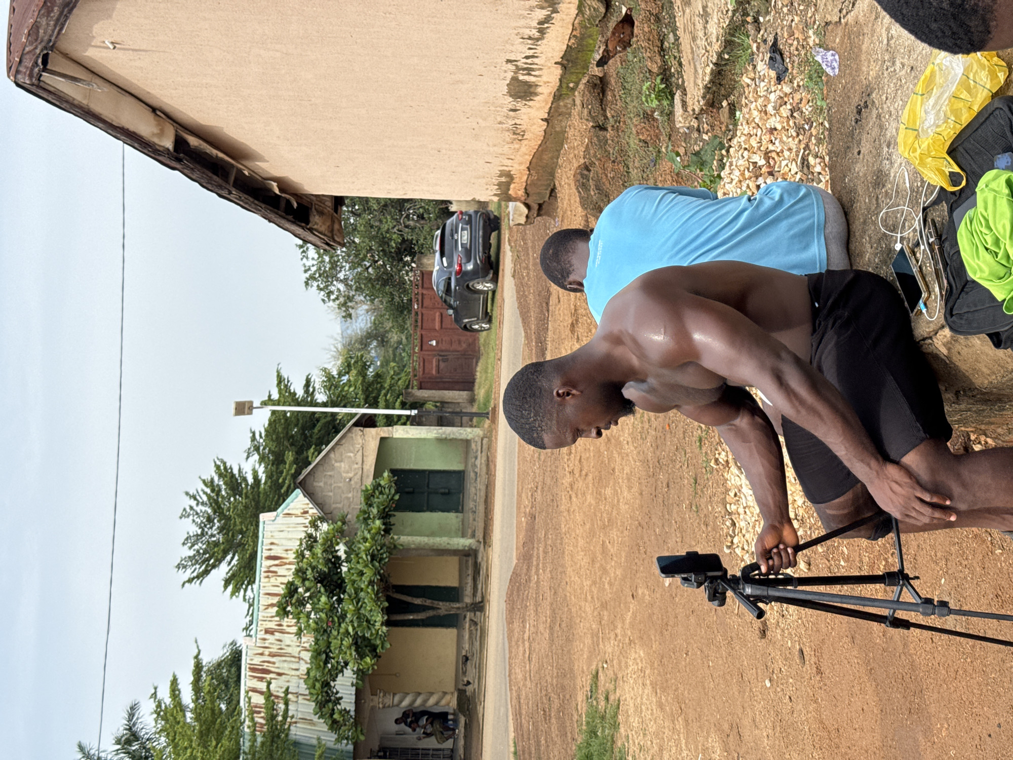 The image depicts an outdoor scene in Akwanga, Nigeria, on a cloudy day. In the foreground, a shirtless Black man with muscular build, wearing black shorts, is seated on a concrete ledge and adjusting a smartphone mounted on a tripod. To his right, another Black person, wearing a light blue t-shirt and grey shorts, is seated facing away from the camera. Scattered on the ground near the concrete ledge are a black bag, a green garment, a yellow plastic bag, wires, and a smartphone. In the midground, a gravelly dirt path leads to a building with a rusted corrugated metal roof, painted in shades of yellow and green with dark green doors. A tree with lush green foliage grows in front of the building. Further in the background, a dark-colored SUV is parked on the right side of the road. Behind the vehicles, a brown wooden fence is visible. The overall environment suggests a somewhat rural or semi-urban setting with visible signs of wear and tear on the structures.