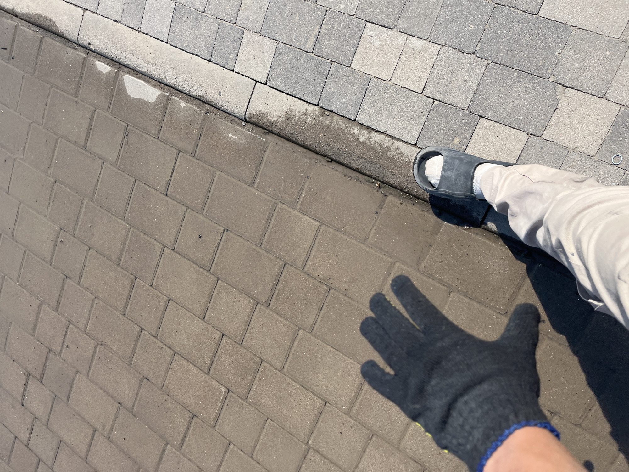 The image shows a close-up of a person's lower legs and feet, as well as a gloved hand, resting on a paved surface. The surface is divided into two sections by a curb, with lighter colored pavers on the upper right and darker, rectangular pavers on the lower left. There are wet patches on the pavers and curb, suggesting recent rain or cleaning. The person is wearing a black sandal on their right foot, a white sock, and light-colored trousers. A black glove with blue trim is visible on the left hand, casting a shadow on the pavers. The scene appears to be outdoors, likely in a park or a residential area given the paving stones. The lighting suggests it is daytime, with the sun casting shadows. The overall impression is casual and practical, possibly indicating yard work or simply a moment of rest.