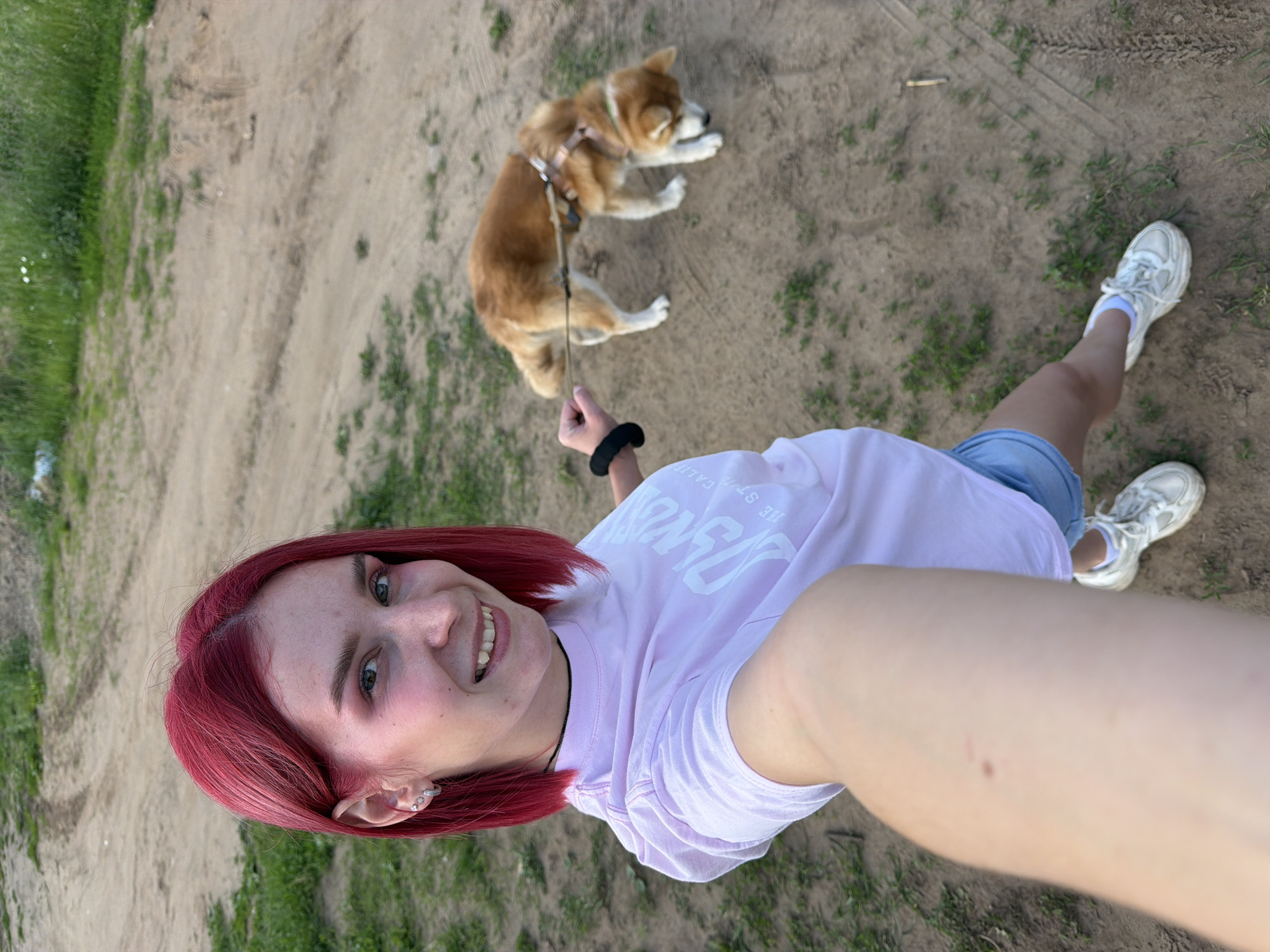 A young Caucasian female with short, bright red hair and fair skin is taking a selfie outdoors. She is smiling and wearing a light pink t-shirt with white lettering and light blue denim shorts. She has on white sneakers and a black choker necklace. Her left hand is holding the leash of a medium-sized, reddish-brown dog that is looking down at the ground. The dog is wearing a harness. They are standing on a dirt path surrounded by sparse green grass and some dry leaves. The background shows more dirt path and distant green foliage. The scene appears to be in a rural or park-like setting, possibly on the outskirts of Chaikovsky, Russia, given the terrain and vegetation.