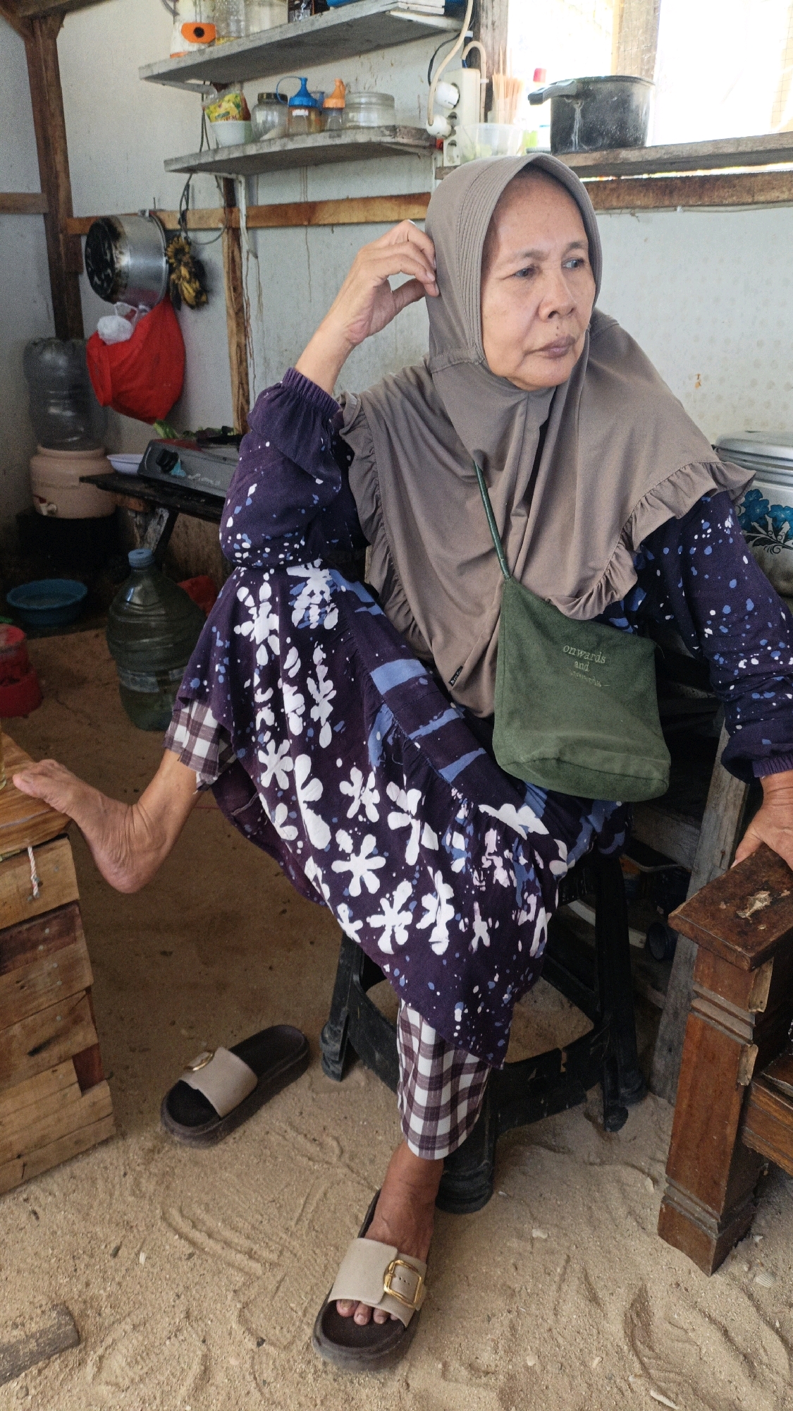 In a room with sandy floors, a woman in a beige hijab and a dark purple, white-floral patterned dress sits on a dark chair. Her left foot, clad in a tan slide sandal, rests on a wooden crate. Another matching sandal lies on the floor near the crate. A small, green crossbody bag with the words "onwards and upwards" is slung across her body. Behind her, shelves hold various containers and kitchenware. A large water jug is visible in the background. The scene appears to be set in the city of Cikal, Indonesia.