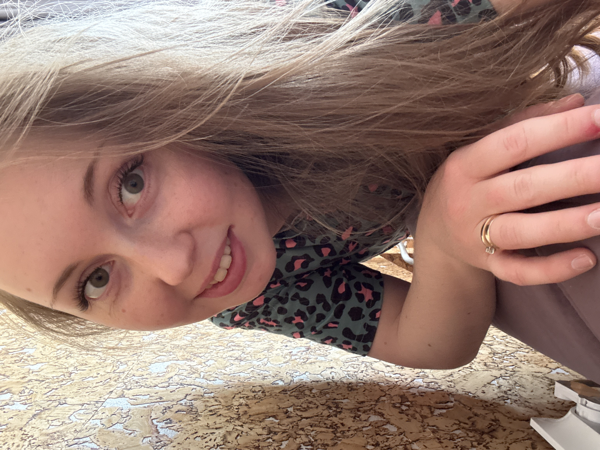 A brightly lit, low-angle selfie captures a young, fair-skinned individual with long, light brown and blonde hair flowing around their face. The person is smiling broadly, revealing white teeth, and looking directly into the camera with light-colored (likely green or hazel) eyes. They have distinct eyelashes and a small mole or freckle below their left eye.
The individual is wearing a short-sleeved top with a dark teal or green base adorned with a repeating pink or coral leopard print pattern. On their right hand, a golden ring with a small, clear stone or open design is worn on the ring finger. A faint reddish mark or scrape is visible on their right index finger, and their fingernails are natural and unpainted.
The background, which occupies the lower and right portions of the frame, appears to be a textured, light brown or golden surface. This surface features an intricate, irregular pattern resembling cracked earth, speckled linoleum, or a decorative countertop. Part of a light-colored, possibly white, object is subtly visible in the far lower right corner. The overall scene suggests a casual and cheerful moment. There is no visible text in the image.