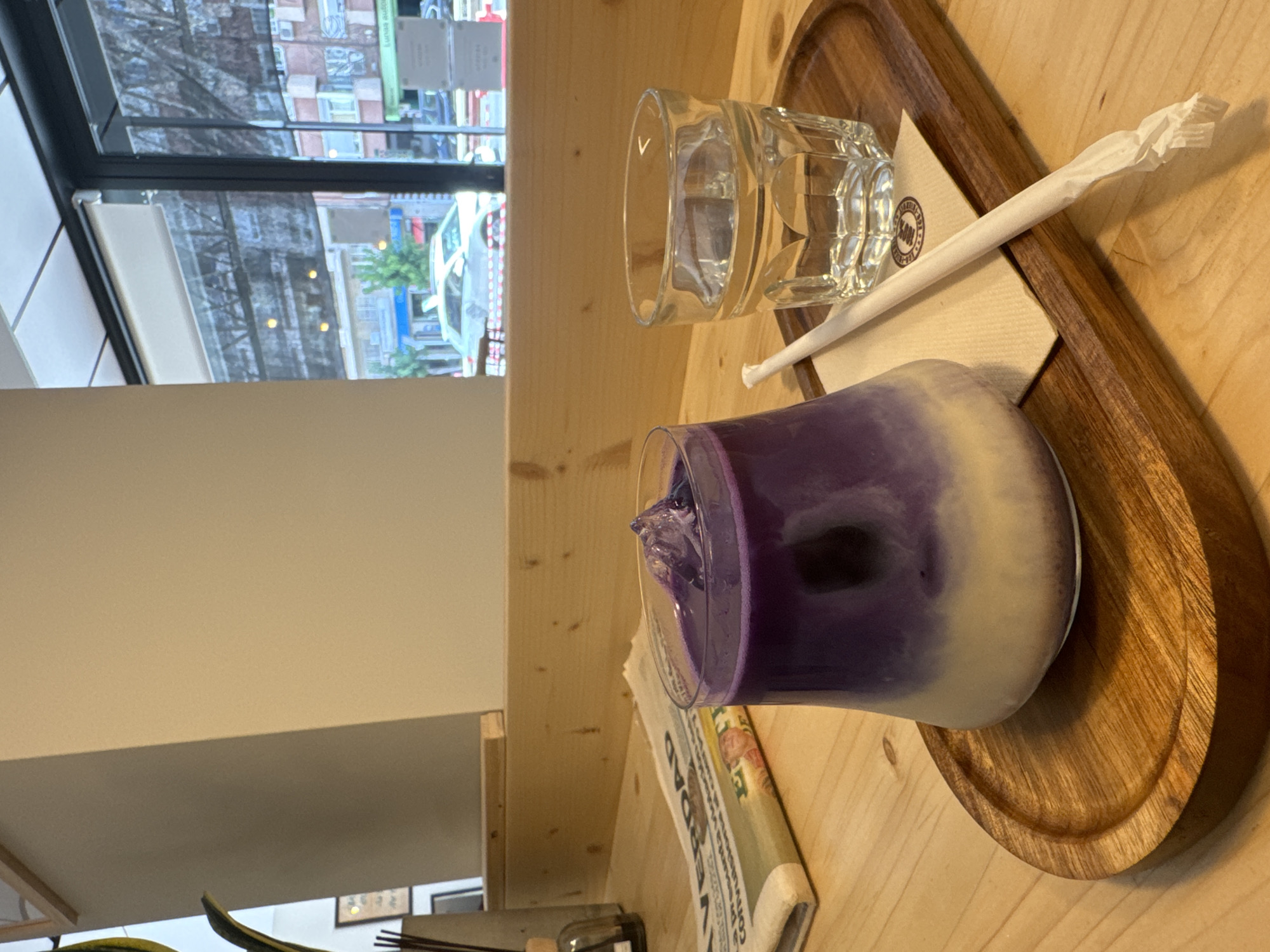 The image depicts a close-up view of a wooden table in what appears to be a cafe in Murcia, Spain. On the table, a glass contains a layered purple and white beverage with ice cubes. Next to it, a second glass is filled with clear water, accompanied by a paper straw resting on a napkin. A folded newspaper lies to the left of the drinks. In the background, through a large window, a street scene with buildings, a car, and trees is visible, suggesting an urban environment. The overall lighting is natural and ambient.