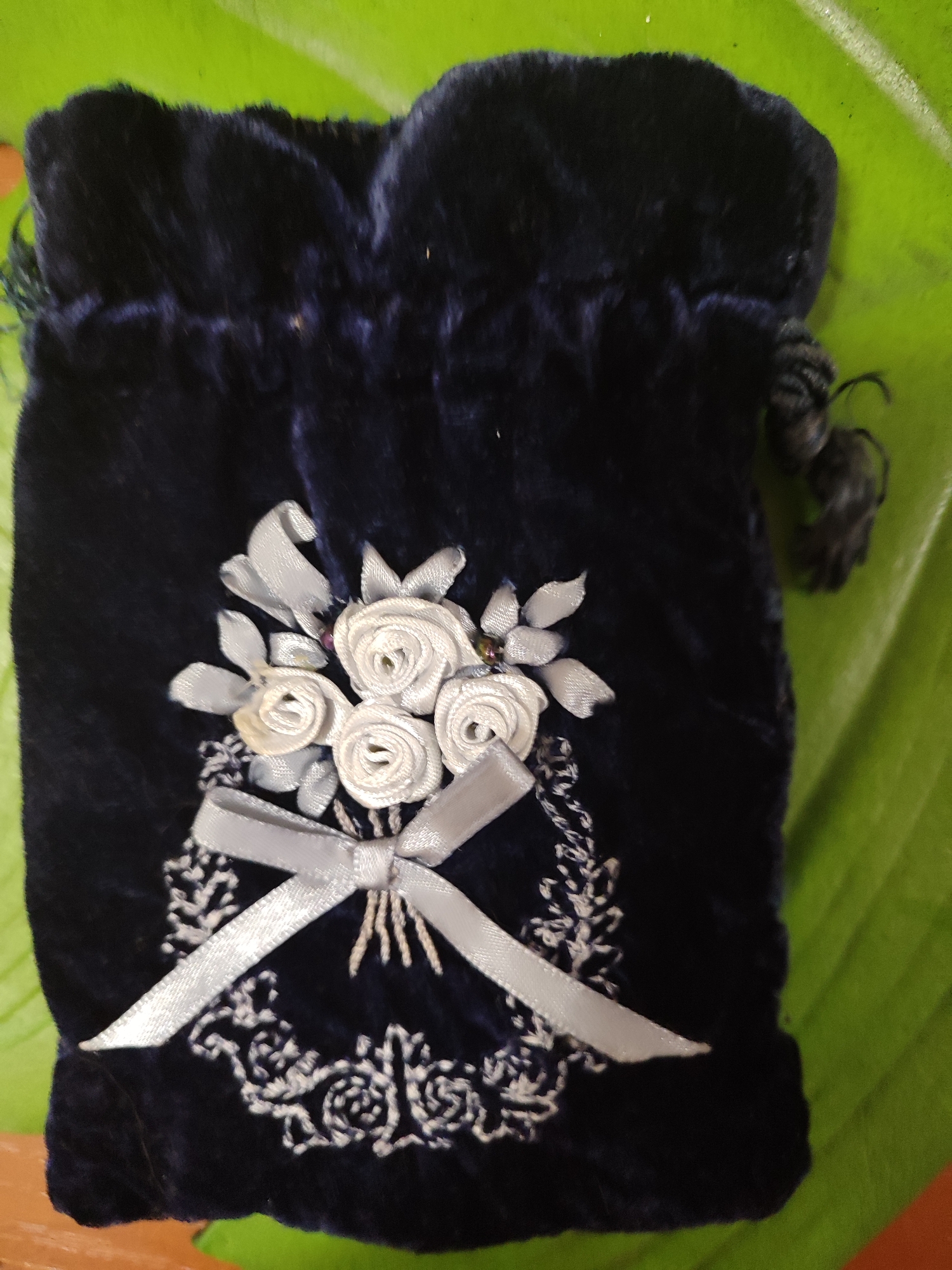 A dark blue velvet pouch, adorned with white ribbon roses and embroidered white floral patterns, rests on a bright green surface. A light grey ribbon is tied in a bow near the center of the pouch. The pouch has a drawstring closure with dark tassels. The background is a textured green surface, possibly a large leaf or fabric. The image is a close-up, top-down view of the pouch. There are no people or identifiable landmarks visible. The scene does not appear to be located in Mykolaiv, Ukraine.