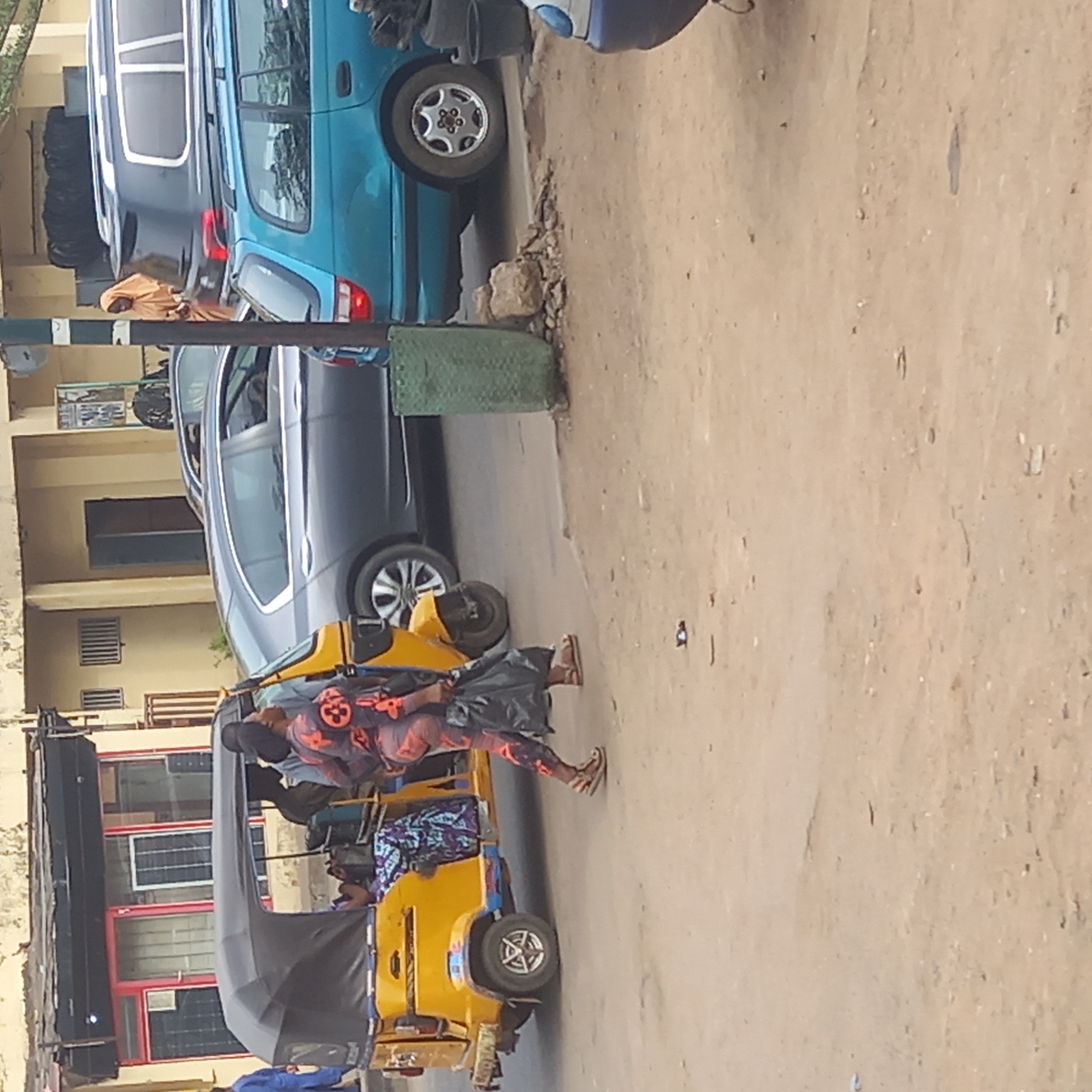 The image depicts a street scene in Dadi Riba, Nigeria. A yellow three-wheeled vehicle (auto rickshaw or "keke") is positioned on the left side of the frame, with passengers inside. A woman is seen walking beside the vehicle. In the background, there are parked cars and buildings. The road appears unpaved.