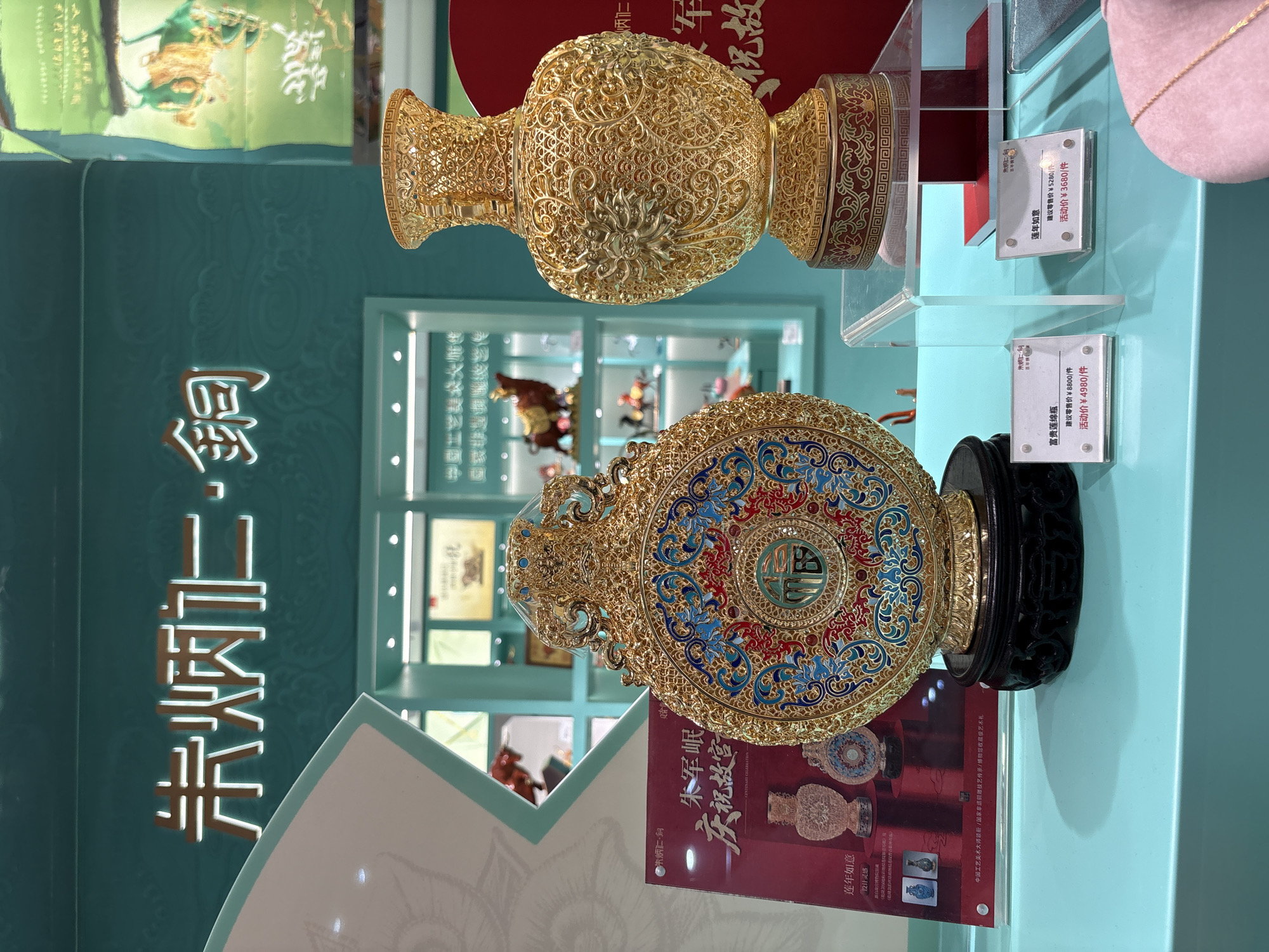 The image shows an indoor display of elaborate gold-plated decorative items, likely in a retail setting in Shanghai, China, based on the Chinese text visible on signage and products. In the foreground, two prominent pieces are displayed on a light blue surface. To the left is a large, circular, ornate object with intricate blue, red, and gold enamel work, featuring a central circular design with Chinese characters that appear to be "福" (fú, meaning fortune). This piece rests on a dark carved wooden base. To the right is a tall, gold-plated vase with a highly detailed filigree pattern covering its entire surface, sitting on a circular red and gold base. Both items are illuminated by overhead lighting, creating reflections on their metallic surfaces.
Behind the foreground display, there are shelves and displays exhibiting more decorative items, including what appears to be a golden ox figurine and other small statues. The wall behind the display features large, illuminated Chinese characters in a stylized font, which read "朱炳仁·铜" (Zhū Bǐngrén · Tóng), indicating the artist or brand and the medium (bronze). Further signage and product information cards with Chinese text are visible, some with prices such as "建议零售价 ¥8800/件" (suggested retail price ¥8800/piece) and "活动价 ¥4980/件" (promotional price ¥4980/piece). The overall environment suggests a high-end shop specializing in artistic metalwork or collectibles, with a clean and well-lit presentation. No people are visible in the image.