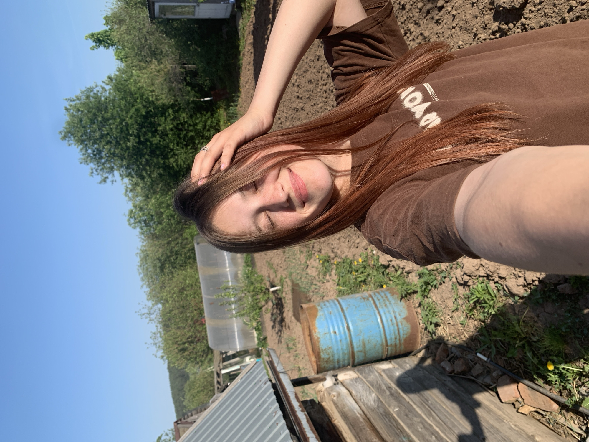 A young woman is taking a selfie outdoors. She is wearing a brown shirt and has long, reddish-brown hair. Her hand is placed on her head.
The background shows a garden or agricultural area. A blue metal barrel sits near a wooden structure, possibly a fence or shed. A greenhouse-like structure is visible further back.
The sky is blue, suggesting a clear day. The location is consistent with a rural or semi-rural setting, potentially in or near Фоки, Russia. The presence of cultivated soil suggests gardening or farming activity.