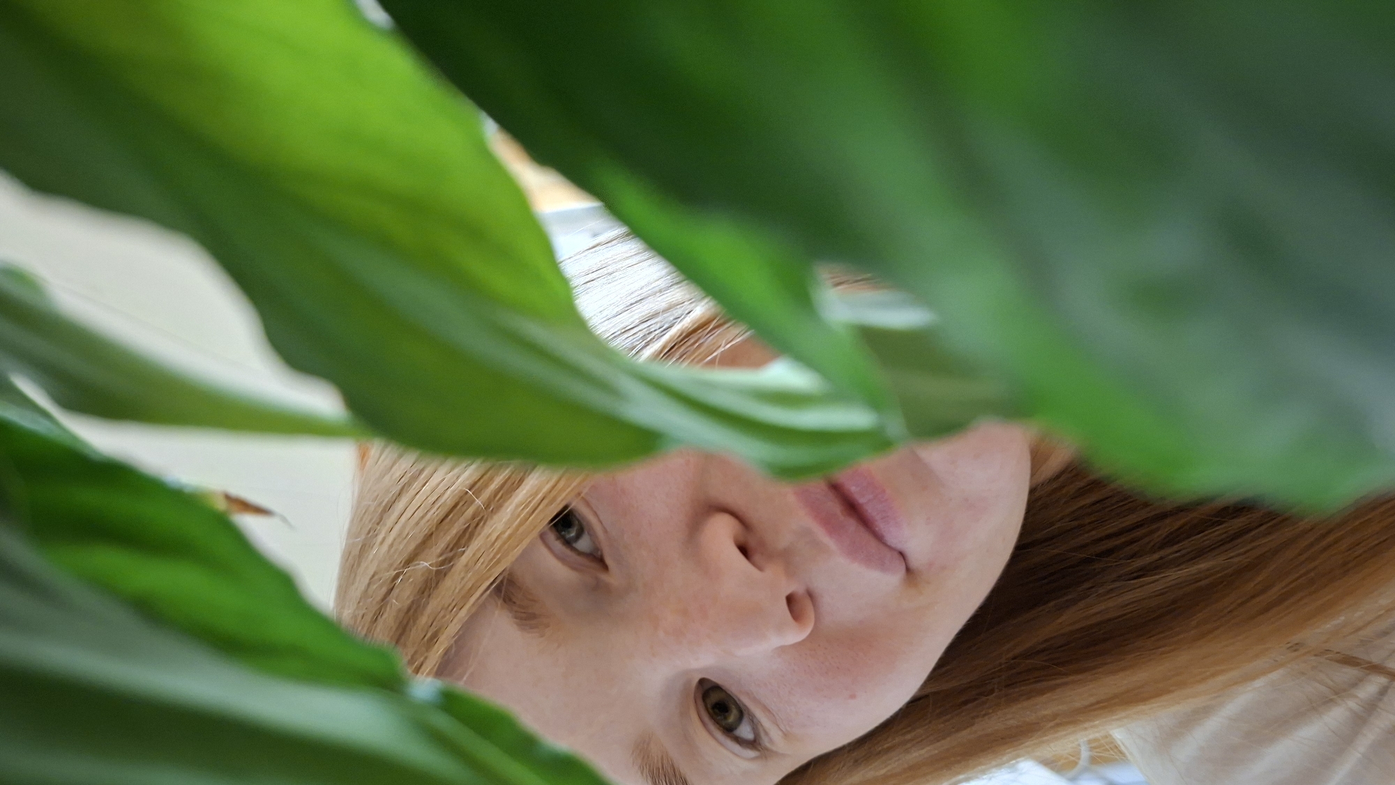 A low-angle, close-up shot captures a young woman with auburn hair peeking through large green leaves. Her face is angled towards the camera, with her right eye visible and looking slightly to the left. She has fair skin with freckles scattered across her cheeks and nose. Her lips are slightly parted and appear to have a light pink lipstick or natural coloring. The leaves in the foreground are blurred and dominate the frame, with their vibrant green color creating a natural framing effect. The background is out of focus, but appears to be an indoor setting with bright, diffused light. The image does not provide any information about the location in the city of Большой Букор, Russia.