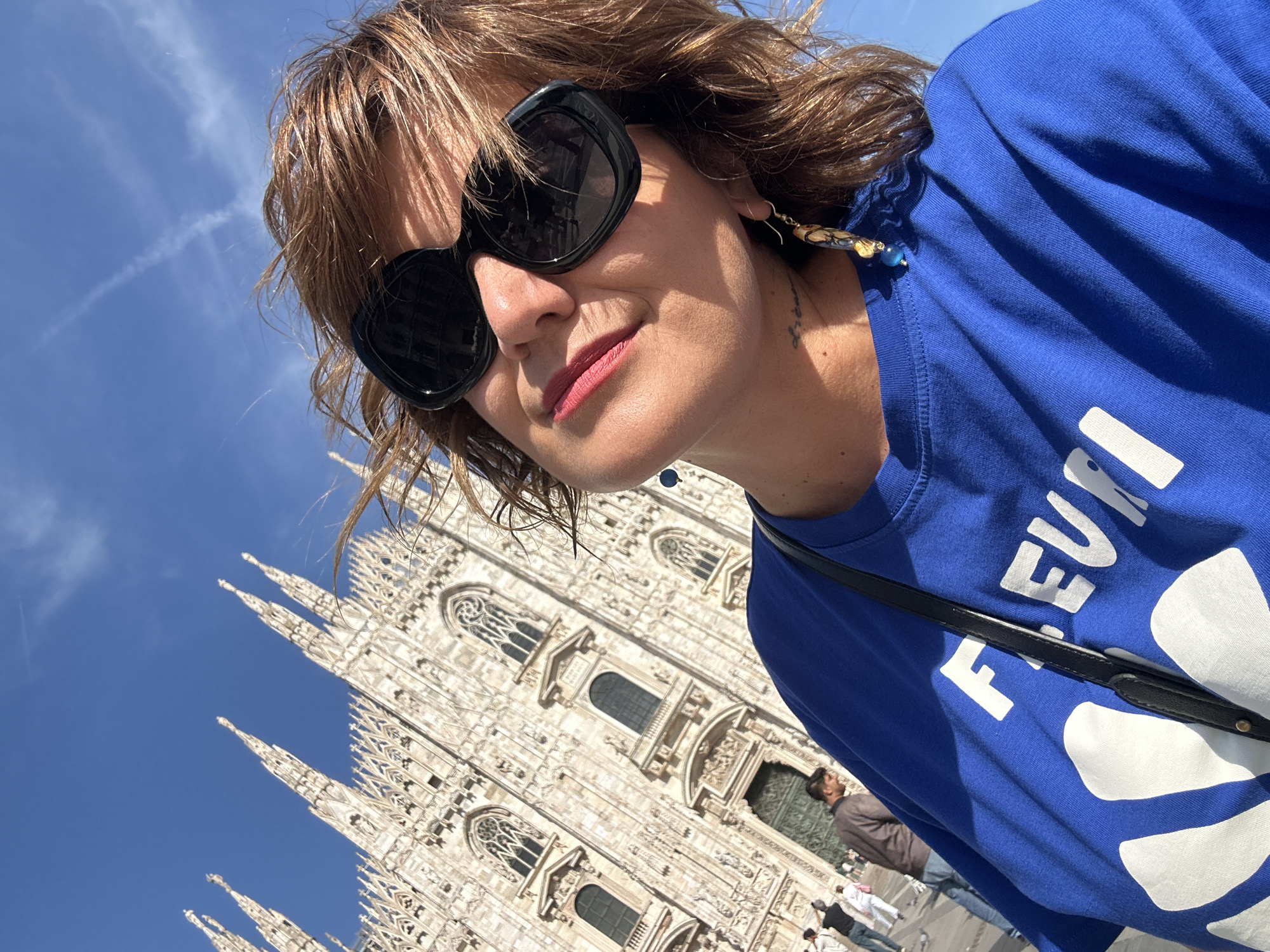 A woman takes a selfie in front of the Milan Cathedral in Milan, Italy. She has brown hair, is wearing sunglasses, and a blue t-shirt with white text on it. She is also wearing earrings and a necklace with blue beads. The Milan Cathedral is a large, ornate Gothic cathedral with many spires. The sky is blue and there are a few clouds. There are other people in the background, walking around.