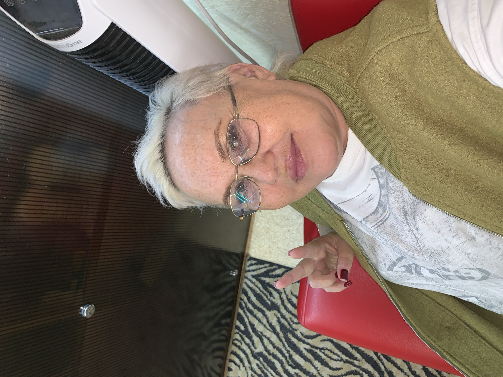 A medium close-up, slightly tilted portrait captures an older woman looking directly at the camera from an indoor setting in Chaikovsky, Russia. She has short, white-blonde hair and fair skin with visible freckles. She is wearing gold-framed eyeglasses and has a subtle smile.
The woman is dressed in a white t-shirt featuring a light grey graphic design, covered by an open, textured olive-green jacket or cardigan. Her left hand is visible, positioned near her waist, with fingers forming a peace sign (V-gesture). Her fingernails are painted with a dark red polish.
She is seated on a bright red, upholstered surface, likely a booth or chair. To her right, a black and white zebra-print patterned carpet or rug is visible. To her left, there is a dark brown or black surface with horizontal ridges, resembling a table or counter. In the upper left background, part of a white and black appliance, possibly an air purifier or fan, is visible, bearing the brand name "Hometome."
The lighting is consistent with an indoor environment. The woman's expression appears calm and friendly, conveying a positive demeanor.