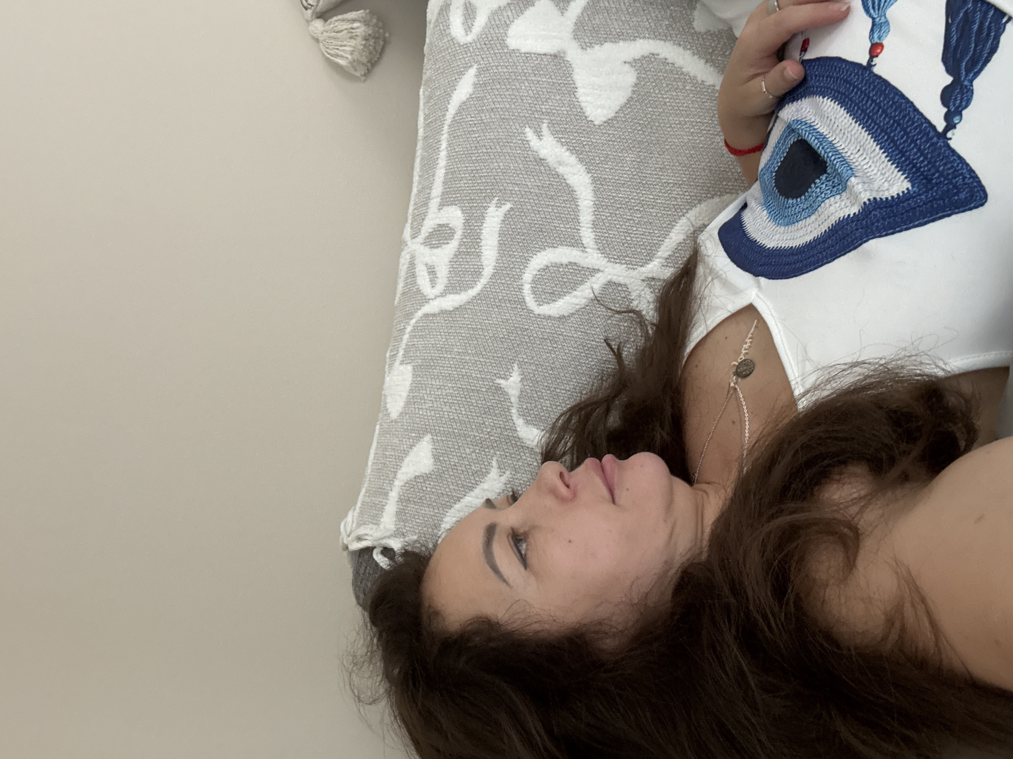 This media file features a person lying down on a bed or sofa, resting their head on a patterned pillow. The pillow is light gray with a white swirling pattern. The person has long, dark brown hair that is spread out around their head. They are wearing a white top that has a prominent blue and white evil eye design on it, along with tassels. A delicate silver necklace with a circular pendant is visible on their neck. A red string bracelet is worn on the wrist. The person's face is visible in profile, with closed eyes. The overall impression is one of relaxation. The lighting suggests it could be daytime, perhaps indoors in a well-lit room. No visible text is present. The scene appears to be set in a domestic environment.