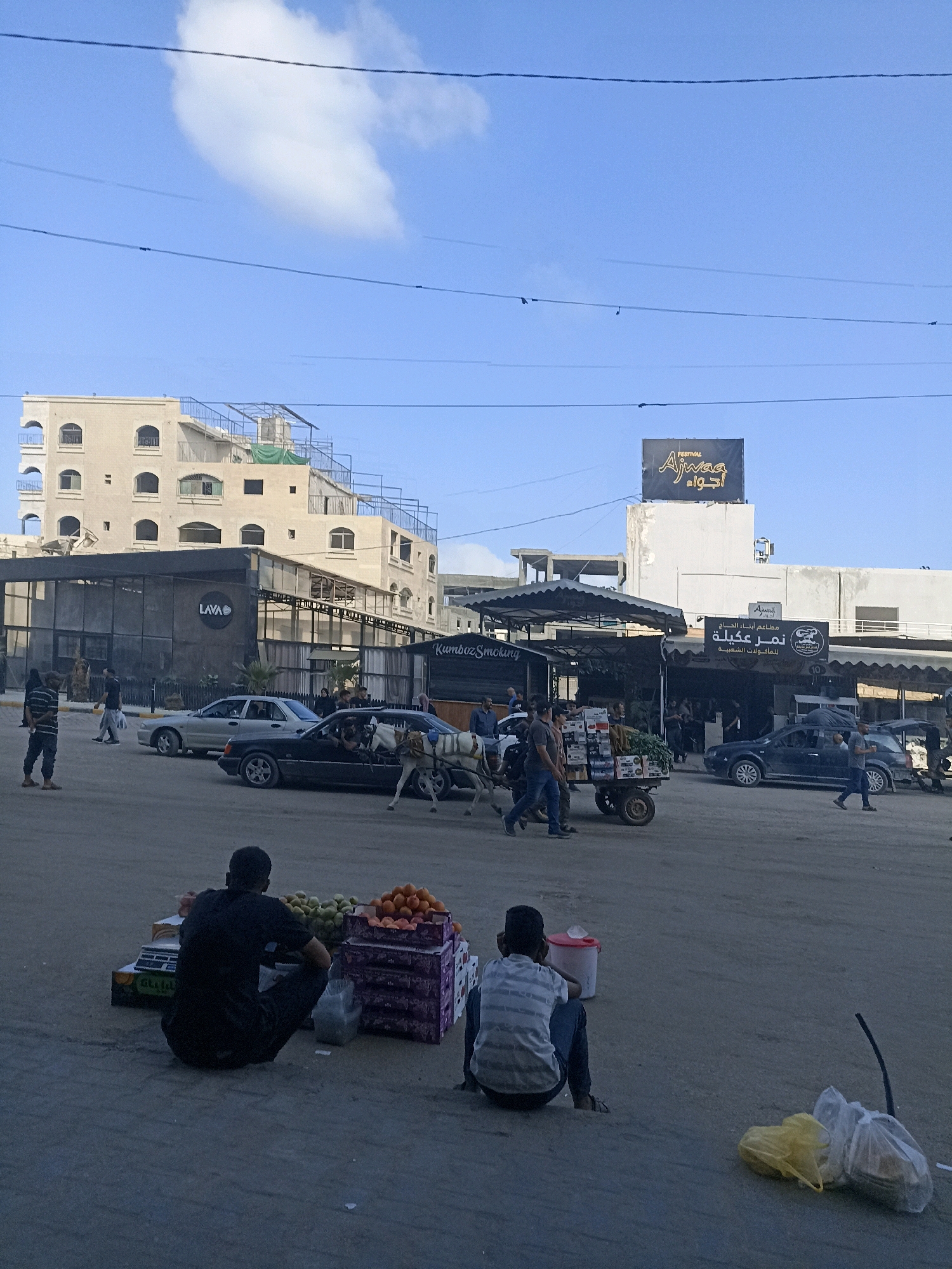 The image depicts a street scene in the city of Gaza, Palestinian Territories. In the foreground, two individuals are seated on the ground near piles of fruit, appearing to be vendors. One person is wearing a black shirt and dark pants, while the other is in a striped shirt and blue jeans. In the middle ground, several cars are parked or driving on the street. A horse-drawn cart laden with goods is also visible, moving across the scene. Various shops and buildings line the street, including one with a sign that reads "LAVA" and another with Arabic script. The background features a multi-story building under construction. The sky above is blue with a few scattered clouds. The overall impression is of a bustling urban environment.