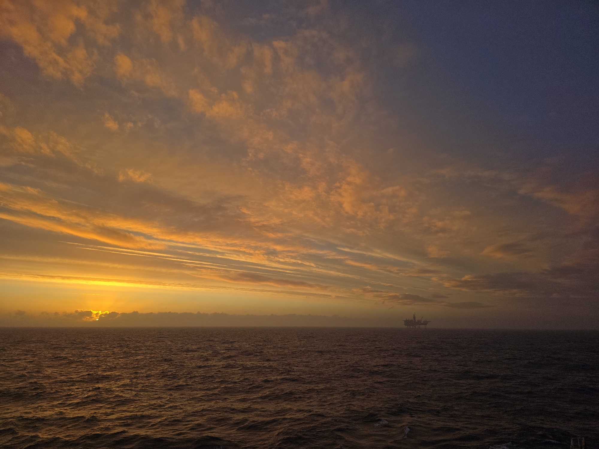 The image depicts a sunset over the ocean. The sky is predominantly filled with orange and yellow clouds, transitioning to a darker blue at the top. The sun is partially obscured by clouds on the horizon, creating a bright glow. The ocean surface is rippled with small waves, reflecting the orange and yellow hues of the sky. A distant offshore oil rig is visible on the horizon. The location is unknown.