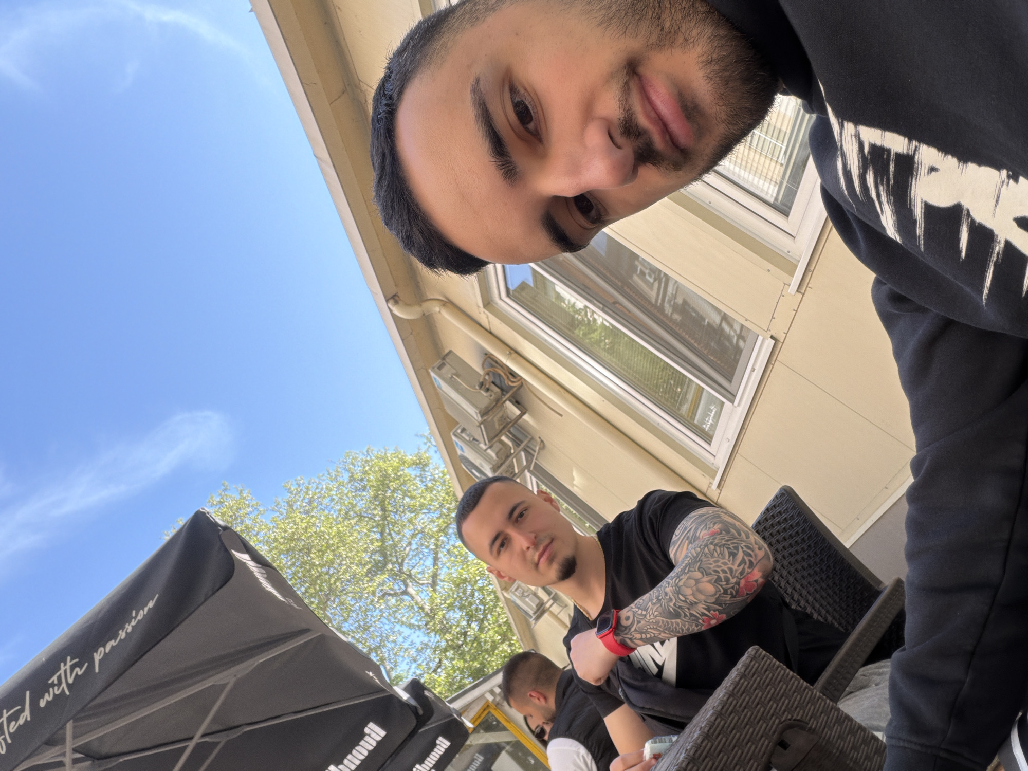 This outdoor shot, likely a selfie taken from a low angle, captures two men relaxing at a café in Plovdiv, Bulgaria, on a clear, sunny day. The man in the immediate foreground, wearing a dark hooded sweatshirt with a white graphic on the sleeve, looks directly at the camera with a neutral expression. His head and shoulders fill the upper right corner of the frame.
Further back, another man is seated at a dark wicker-style table, also looking towards the camera. He has short dark hair, a goatee, and prominent full-sleeve tattoos on his left arm. He wears a black t-shirt, possibly with a white "NI" logo, and a red watch on his left wrist. A third person is partially visible behind him. The setting suggests an outdoor dining area, possibly part of a restaurant or bar.
Notable objects include dark wicker chairs and tables, and a large black outdoor umbrella. Part of the text "ted with passion" is visible on the umbrella, along with an inverted fragment "hnoudi". In the background, a building with light-colored siding and windows is visible, along with an external unit (possibly air conditioning) mounted on its wall. Lush green trees and a bright blue sky with a few wispy clouds indicate pleasant weather, likely midday or afternoon. The overall mood is casual and relaxed.