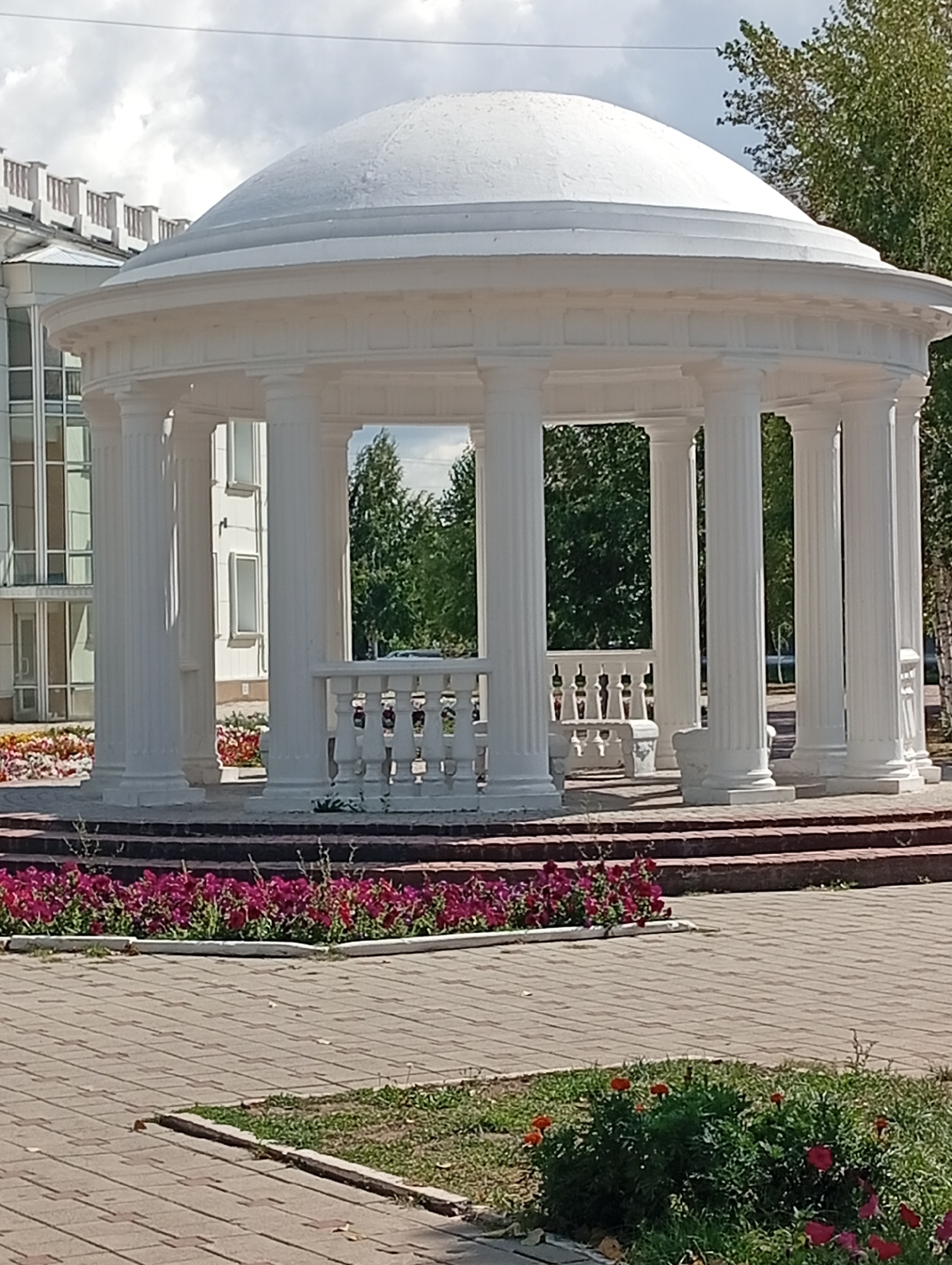 The image depicts a white, neoclassical rotunda with a domed roof, supported by fluted columns. A balustrade surrounds the interior, with a stone bench visible inside. The rotunda is situated in a park-like setting, with a paved walkway in the foreground and a flower bed of purple flowers to the left. In the background, a modern building with large windows can be seen, along with trees and a cloudy sky. This rotunda is located at the entrance to the Kostanay Regional Museum of History and Local Lore in Kostanay, Kazakhstan.