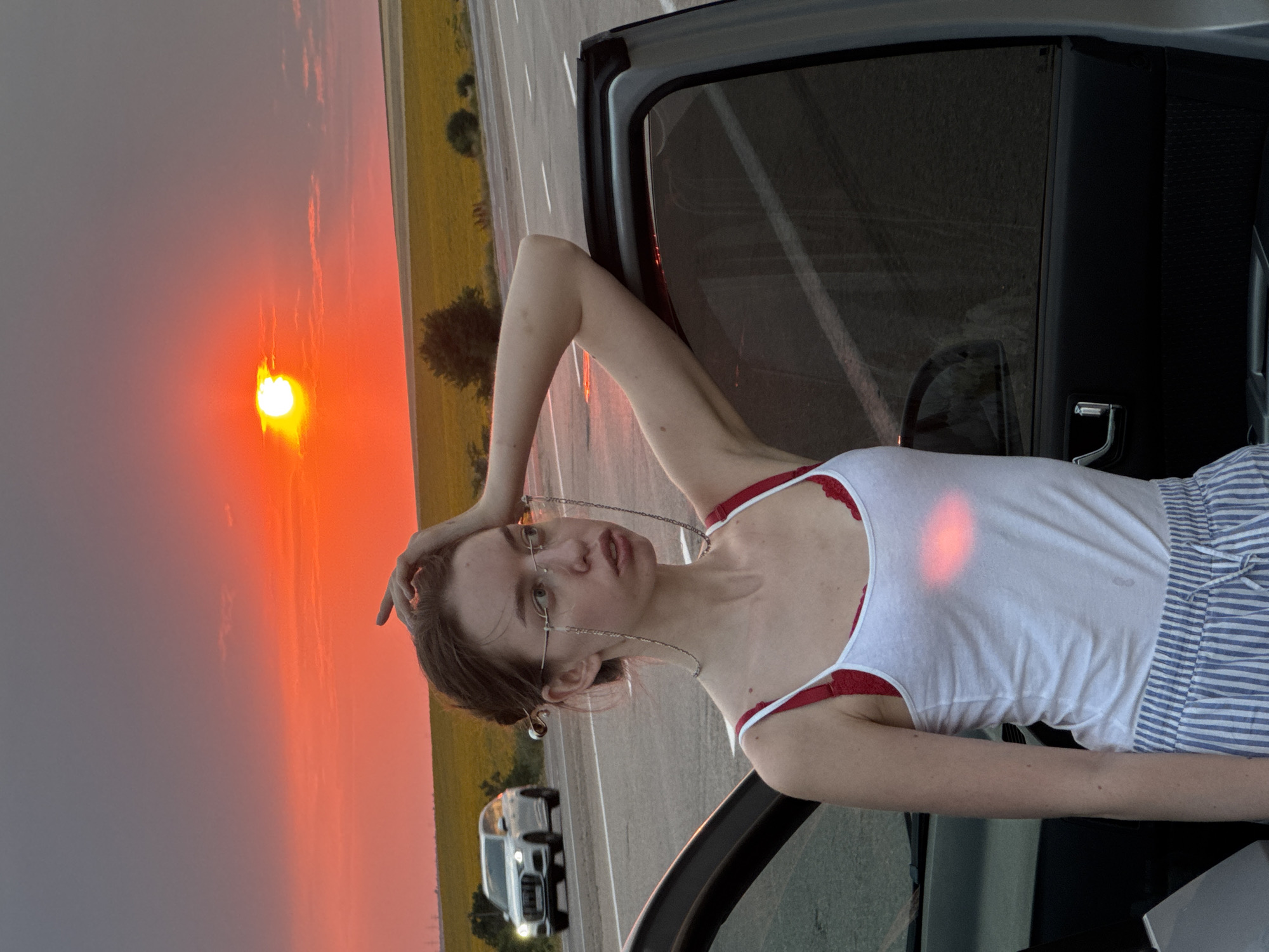 A young woman with brown hair pulled back, wearing glasses with chains, a white tank top, and striped shorts, leans against the open door of a car. The scene is set by a road with a field of yellow flowers in the background, bathed in the dramatic orange and red light of a setting sun. A white SUV is visible driving on the road in the distance. The location is Yelyzavetivka, Ukraine.