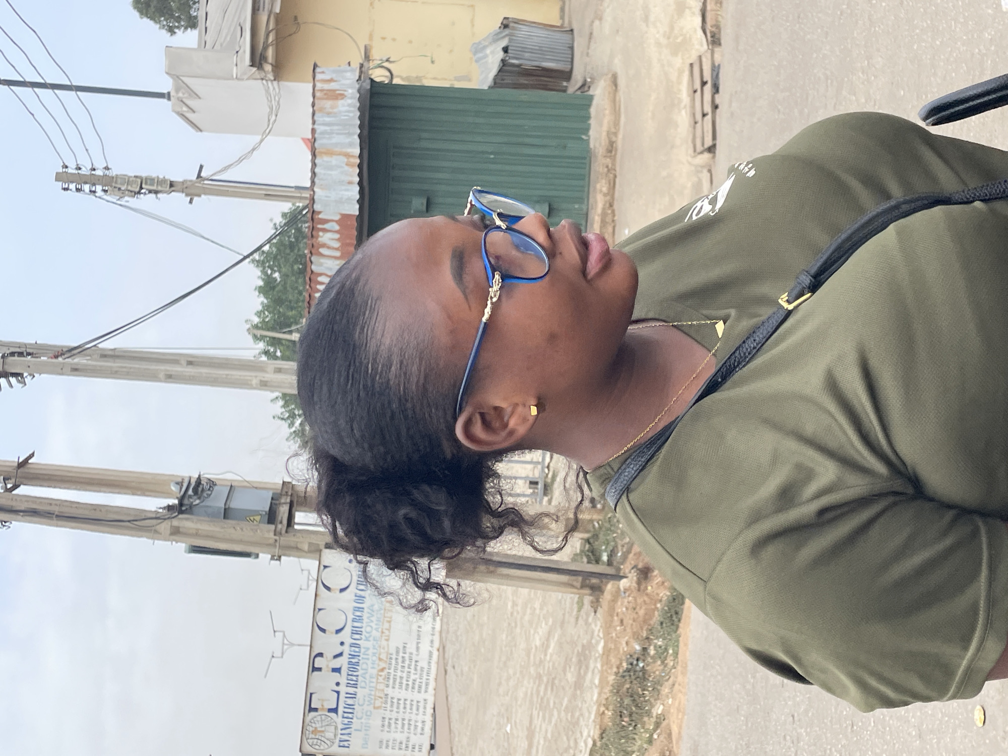 The image shows a woman wearing glasses, an olive green shirt, and a black shoulder bag, captured in Jos, Nigeria. In the background, an Evangelical Reformed Church sign is visible, reading "E.R.C C EVANGELICAL REFORMED CHURCH OF CH L.C.C. DADIN KOWA BEHIND WHITE HOUSE ANGVA". Overhead electrical lines and utility poles are present. Behind her the surroundings include corrugated metal buildings and foliage.