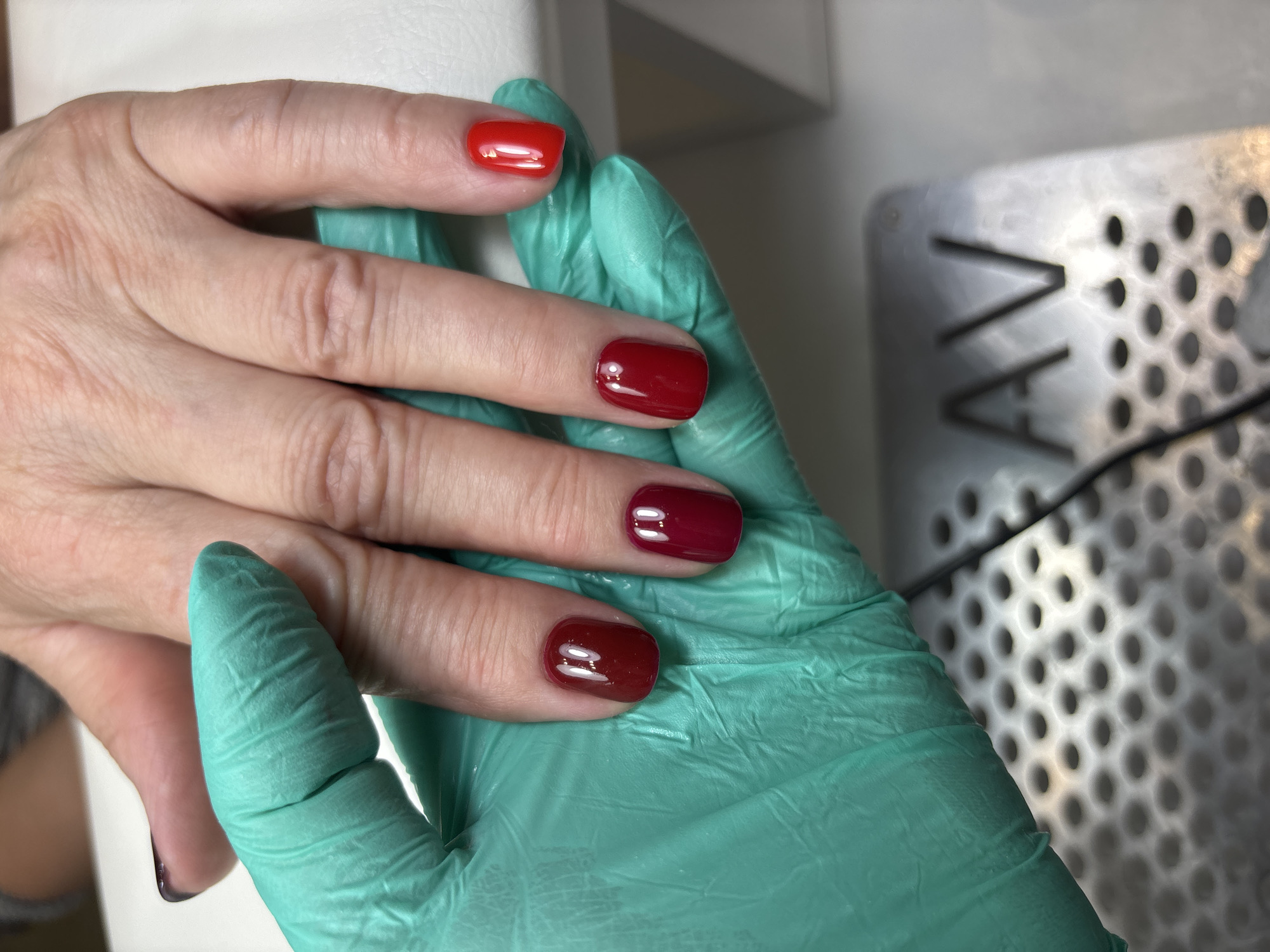 A close-up photograph depicts a person's left hand adorned with varying shades of red nail polish. The index finger exhibits a bright, classic red, while the remaining fingers showcase deeper, richer tones of red. The hand is resting on a light green glove, presumably worn by a professional or caregiver. The background includes a white surface and a metallic object with a perforated design. The location in Ферма, Russia is not evident from the image.