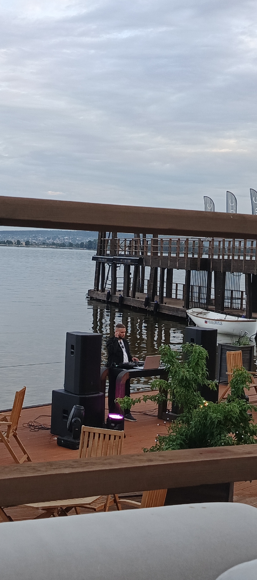 A man is standing at a DJ booth on a wooden deck by a body of water. He is wearing a black suit and appears to be a DJ, with a laptop and sound equipment in front of him. There are several large black speakers stacked next to him, and a purple stage light on the ground. Wooden chairs are placed around the deck. In the background, a wooden pier extends into the water, with a white boat moored to it. A town with buildings and trees is visible on the distant shore. The sky is overcast.