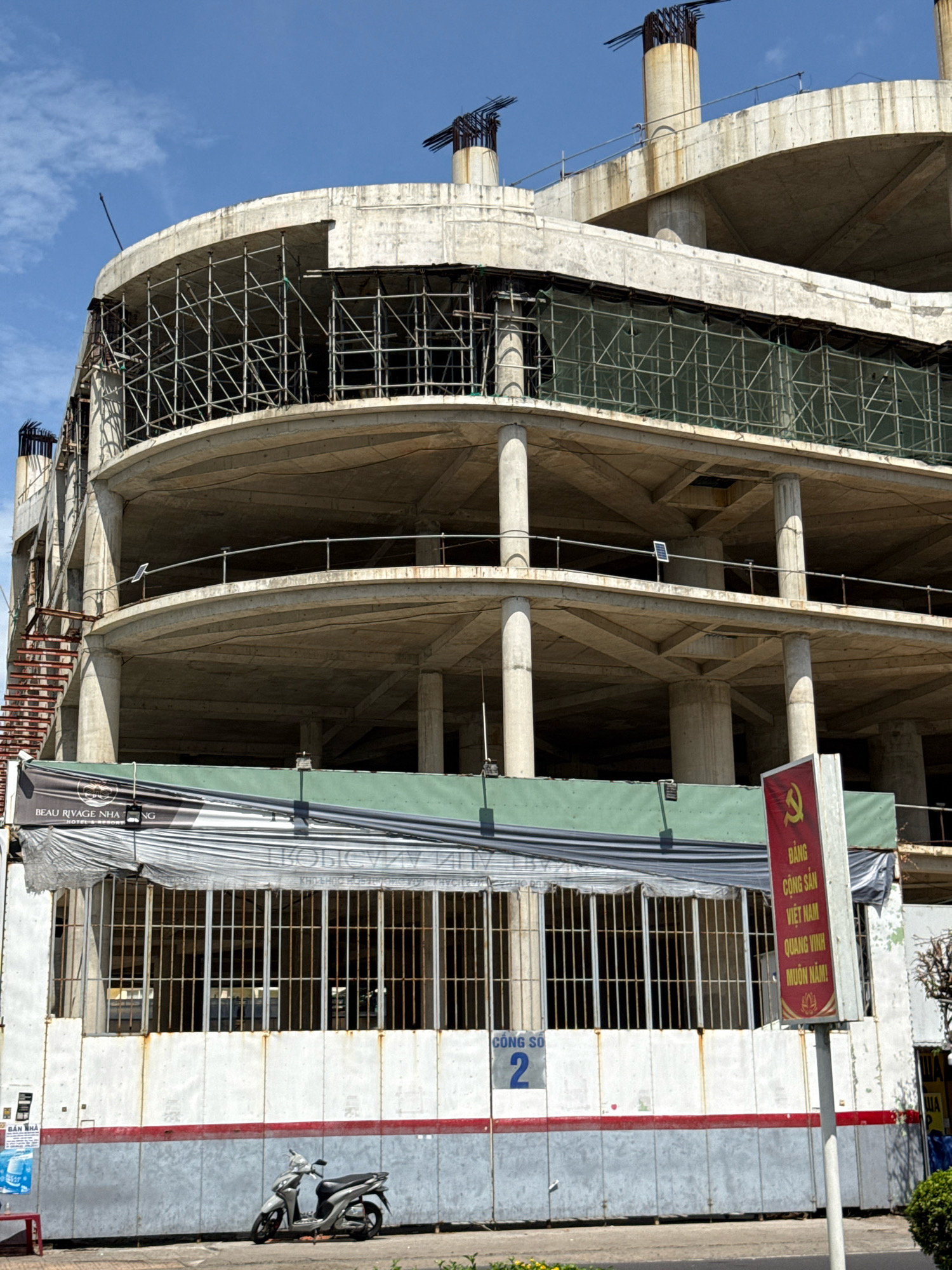 The image shows a partially constructed building with multiple levels and a curved facade, located in Nha Trang, Vietnam. The structure is made of concrete and steel, with scaffolding and green netting visible on the upper floors. Signs with Vietnamese text are attached to the lower part of the building. One sign prominently displays the hammer and sickle symbol and text that reads "ĐẢNG CỘNG SẢN VIỆT NAM QUANG VINH MUÔN NĂM!" which translates to "Long live the Communist Party of Vietnam!". Another sign indicates "CÔNG SỞ 2", meaning "Office 2". A silver scooter is parked on the sidewalk in front of the building. The sky is blue with some clouds.