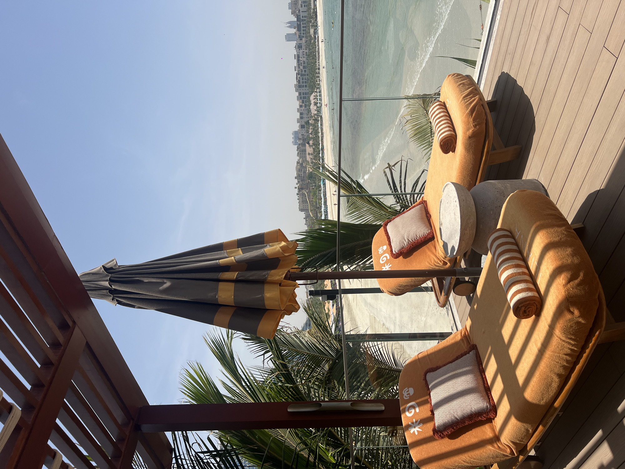 Two lounge chairs, an umbrella, and a small table are on a wooden deck overlooking a beach and the city of Dubai. The lounge chairs are orange with striped bolster pillows and square pillows. The umbrella is closed and striped in gray and gold. A clear glass railing separates the deck from the beach. Palm trees and other greenery are visible. The beach has white sand and turquoise water. The Dubai skyline is visible in the background.