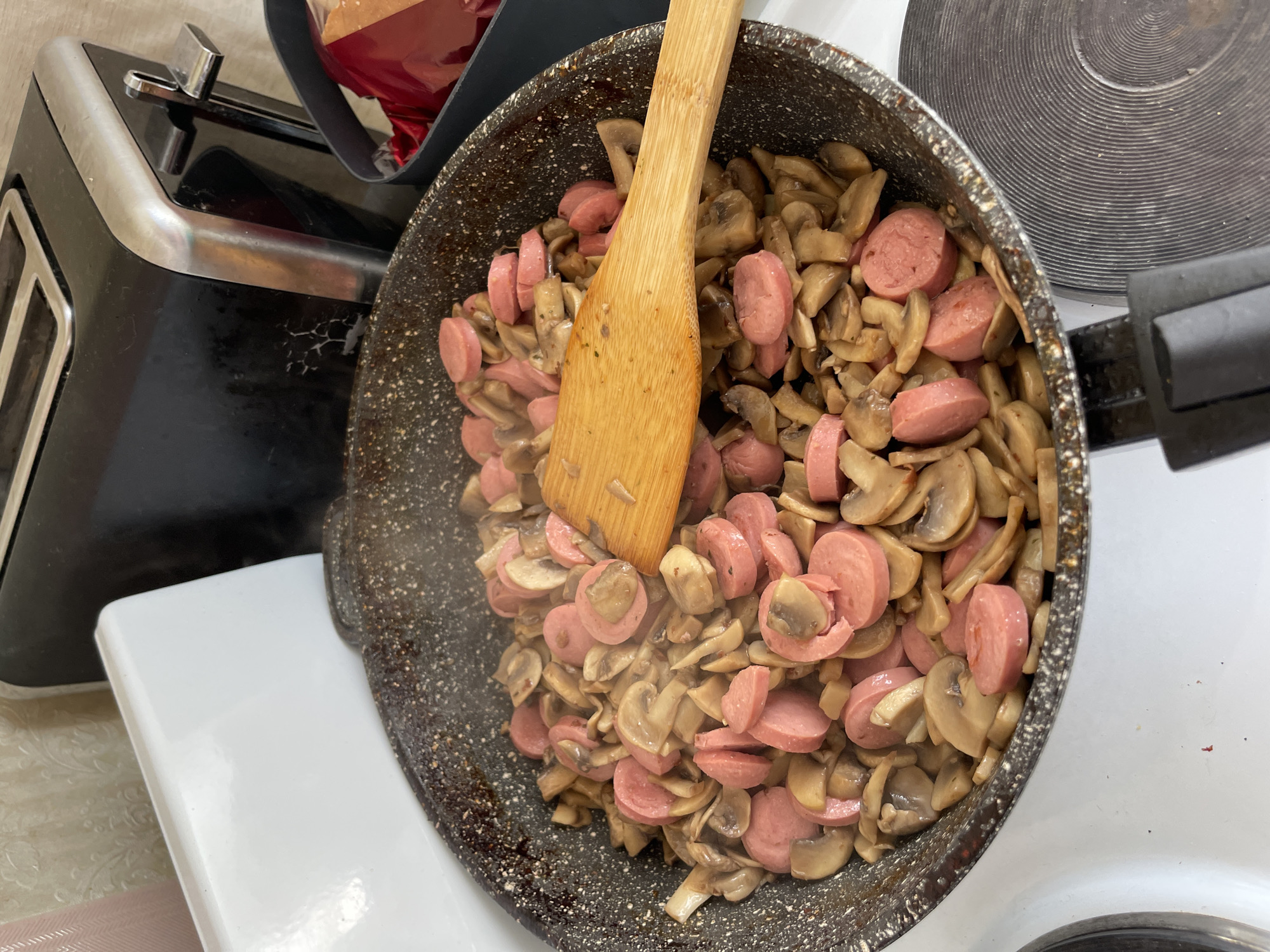 The image shows a close-up view of a frying pan filled with sliced sausages and mushrooms being cooked. A wooden spatula is resting on top of the food, mixing it. The pan is on a white stovetop. In the background, a black toaster is visible, as well as part of a kitchen appliance with a textured, light-colored surface, possibly a backsplash. There's also a dark gray container with red items inside. The overall setting appears to be a domestic kitchen. The image does not provide specific details to determine the location within the city of Khimki, Russia.