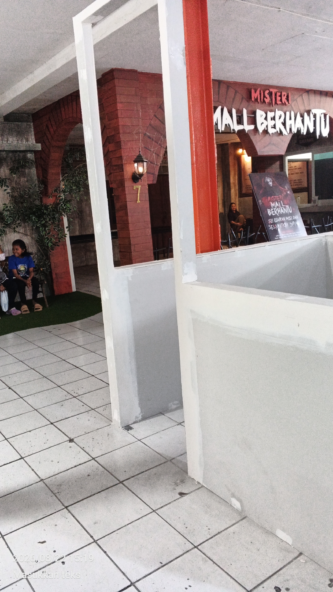 An outdoor shot captured at "Misteri Mall Berhantu" in Malang, Indonesia, shows a festive yet slightly eerie ambiance. The foreground features two young girls, one in a blue shirt, sitting on the ground with an amused expression. In the background, a sign for "Misteri Mall Berhantu" is prominently displayed, adorned with spooky imagery. The mall's facade is decorated with brickwork arches and a rustic lamp, contributing to the haunted theme. A large red pillar stands tall, framing the scene. The tiled floor, a common sight in Indonesian public spaces, shows signs of wear and tear, suggesting a well-trodden path.
