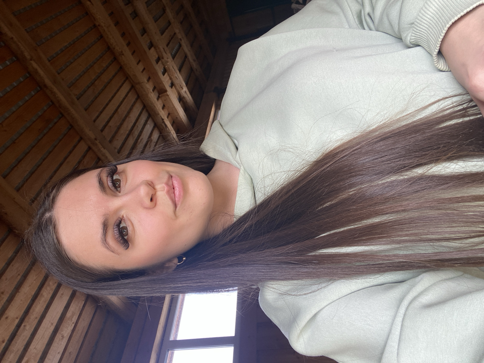 A woman is seen from a low angle, tilted to the right. She has long, straight, dark brown hair and wears a light green hooded sweatshirt. Her makeup includes enhanced eyelashes. The background features a wooden ceiling with linear patterns and a bright window. The location is impossible to determine from this image.