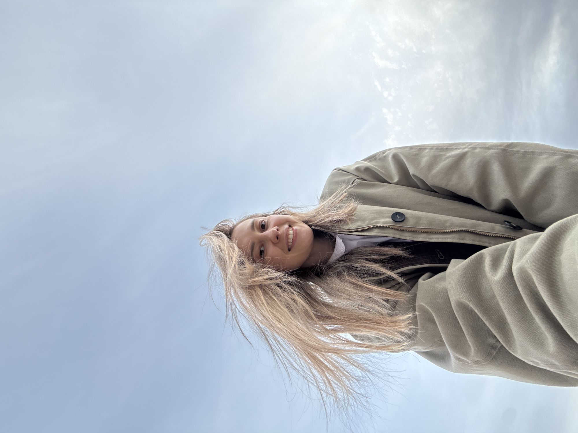 A woman with long, blonde hair smiles at the camera from a low angle. She is wearing a beige jacket and a white shirt. The background is a cloudy sky. The location is Chaikovsky, Russia.
