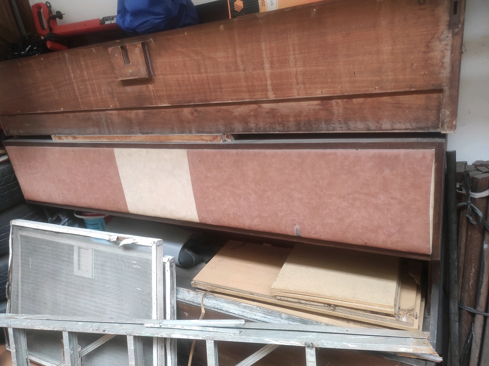 The image shows a storage area in what appears to be a workshop or garage. In the foreground, a metal ladder is visible, supporting a white-framed window screen. Behind the ladder and screen, stacks of particle board or plywood are arranged. The middle ground is dominated by a large, wooden chest or cabinet with a two-toned front panel, featuring a lighter beige stripe down the middle and a darker reddish-brown on the sides. This chest rests on a lower structure, also made of wood. Above the chest, various items are stacked, including what looks like a red power tool, a blue bag, and some cardboard boxes. The background consists of a white wall and a stack of wooden planks to the right, bound with black rope. The lighting suggests an indoor environment, likely with artificial or diffused natural light. There are no people visible in the image, and the scene is static.
