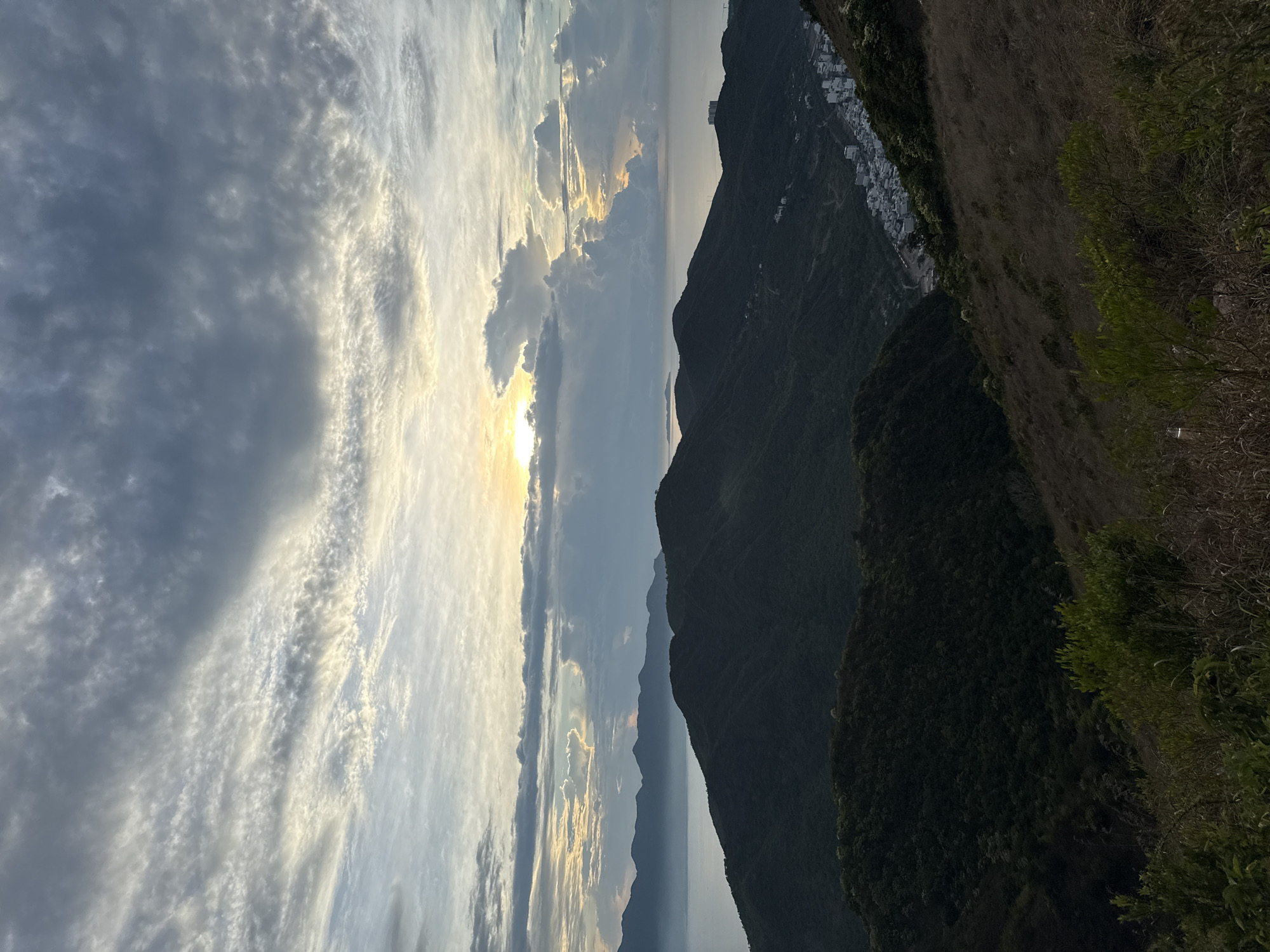 The image shows a mountain landscape at sunset. In the foreground are mountains covered in dark green vegetation and a dirt slope with dry grasses. In the distance, a body of water is visible, partially obscured by mist or haze. Above, the sky is filled with dramatic clouds lit by the setting sun. A line of white buildings clings to the side of one of the distant mountains. The image appears to be taken in a mountainous region of Nha Trang, Vietnam.