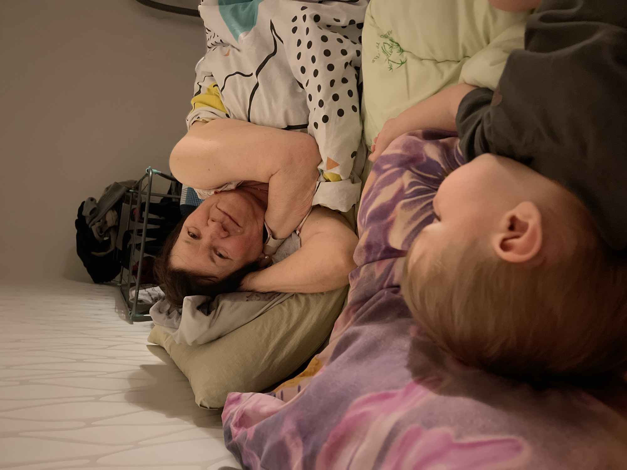 An intimate, sideways-oriented indoor image captures two individuals resting in what appears to be a bedroom in Perm, Russia.
The primary subjects are an adult woman and a young child. The woman, possibly a mother or grandmother, is on the left, lying on her back with her head on a light beige pillow. She has dark hair, is wearing a light-colored top, and looks towards the viewer with a gentle, contented smile. Her left arm is bent and rests near her head. She is partially covered by a white duvet with black polka dots and abstract black lines, with hints of yellow fabric underneath.
To her right, partially obscured and in the foreground, is a young child, likely a baby or toddler. The child is lying face down or on their side, with the back of their light blonde or light brown head clearly visible. They appear to be sleeping peacefully, covered by a purple and pink patterned blanket or duvet. A dark grey top is visible on the child's upper body.
The setting is a domestic bedroom. In the background on the left, a metal shelving unit or clothes rack is visible with various clothes on it. The room has plain white walls. The floor in the foreground on the left features a distinct white pattern with wavy, light grey lines.
The overall activity depicts a quiet moment of rest or sleep, suggesting co-sleeping or sharing a bed. The woman's expression conveys affection or serenity. A light green pillow, possibly belonging to the child, is visible between the two individuals, featuring a faint, unreadable green design or text resembling stylized bamboo. The indoor lighting suggests it could be night or early morning.