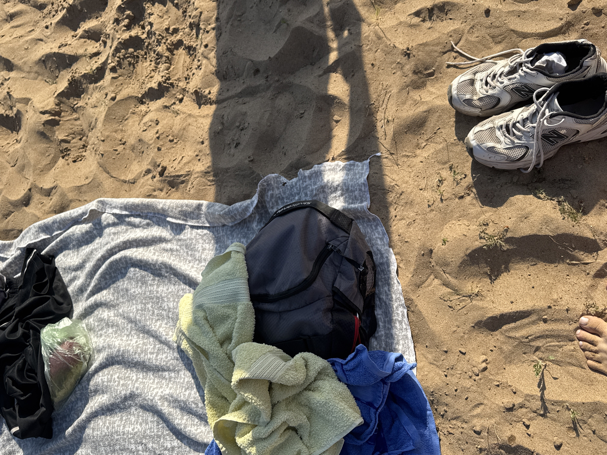 The image depicts a beach scene, presumably in Novyi, Russia, given the prompt's context. The ground is sandy with some scattered vegetation. In the upper right corner, there's a pair of white and grey New Balance running shoes. A shadow is cast on the sand, possibly from the photographer. In the lower portion of the image, a grey patterned towel is spread out. On the towel, there are black shorts, a clear plastic bag containing an unidentifiable item, a backpack, and a yellow and blue towel. A foot is visible in the lower right.