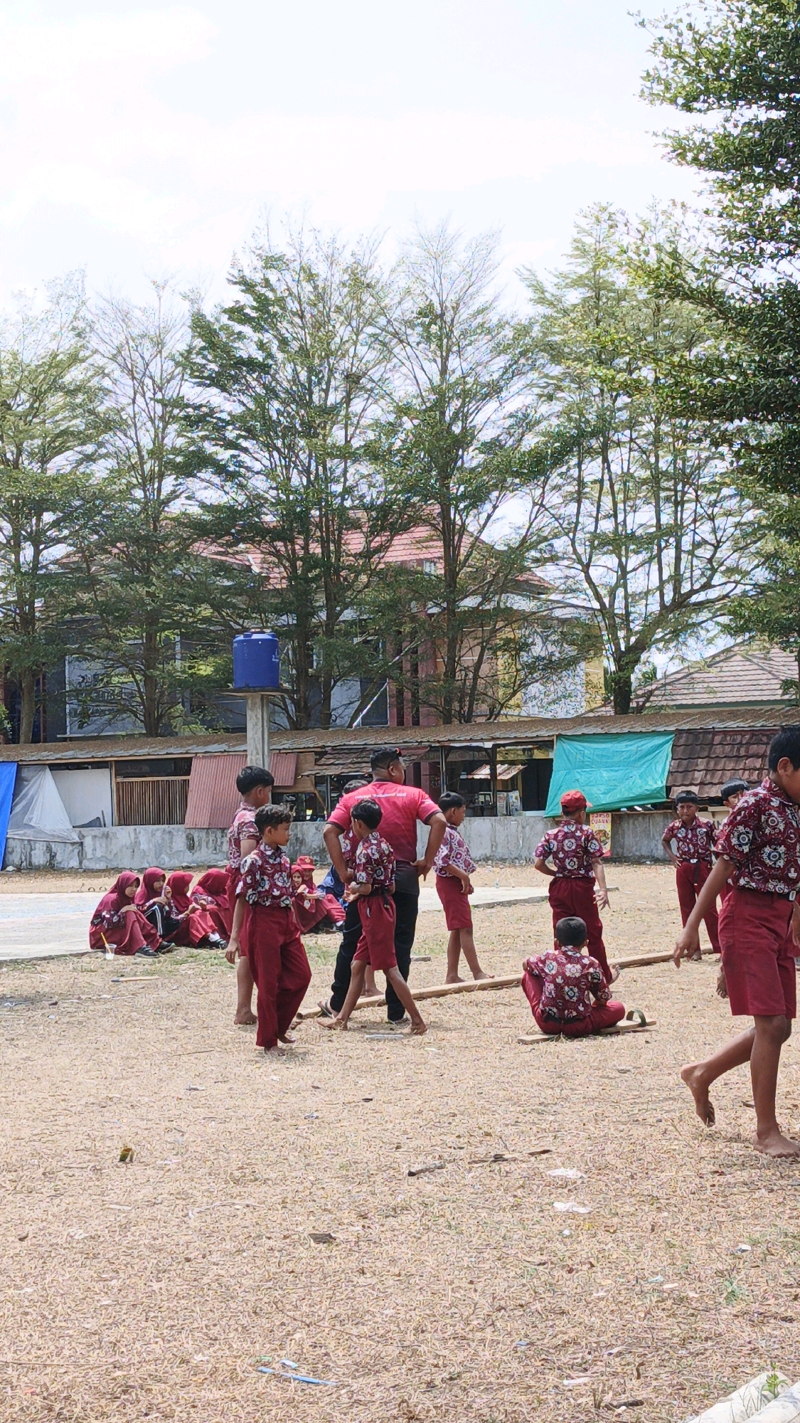 In an outdoor area, likely a schoolyard in Sukaresik, Indonesia, a group of children in matching maroon and white patterned uniforms are engaged in various activities. Several children are seated on the ground, some on a low wall or ledge, while others stand or move around. One adult, wearing a bright red t-shirt and black pants, appears to be supervising the children. The ground is covered in dry, light-brown material, possibly dirt or sand. In the background, there are buildings with tiled roofs and trees with dense green foliage. A blue water tank is visible on a concrete pillar. The sky is overcast.
