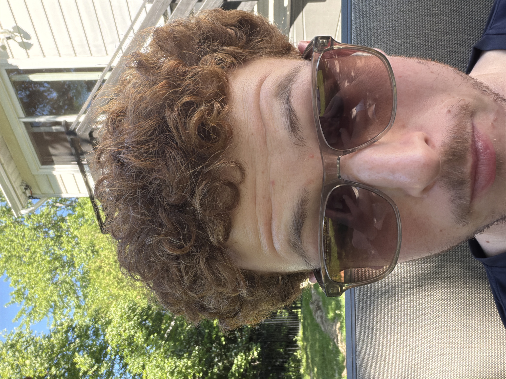 This close-up, horizontally oriented image, taken in Pepper Pike, United States of America, captures a person relaxing outdoors on a sunny day. The subject, visible from the forehead to the chin, has curly, reddish-brown hair and is wearing brown-tinted sunglasses with transparent, slightly greenish-brown frames. The person's eyes appear closed or lowered behind the dark lenses, and several horizontal wrinkles are visible across their forehead. Sparse facial hair (stubble) is present around the chin and upper lip area.
The background elements suggest an outdoor setting. To the left, part of a white-sided building with reflective windows is visible, showing reflections of green trees and blue sky. Lush green trees and a clear blue sky dominate the background to the right and above the person's head. The subject appears to be seated or reclined on textured outdoor furniture, possibly a patio chair, indicated by a woven fabric pattern in shades of gray. A dark blue collar is visible near the bottom right. The bright light and the presence of sunglasses indicate it is daytime under clear, sunny weather conditions. No text is visible in the image.