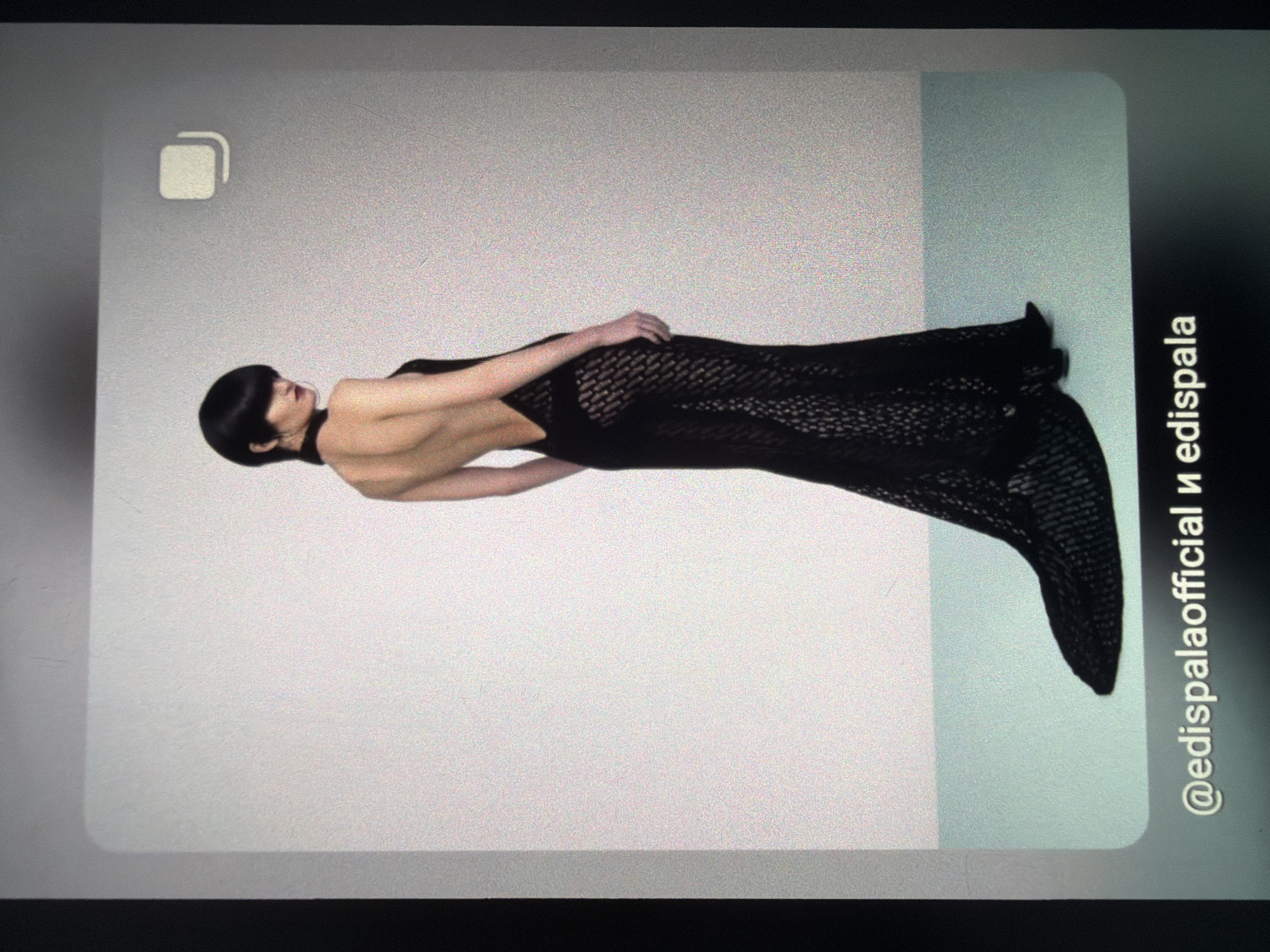 A full-body, side-view photograph shows a model standing with her back to the camera. She is wearing a long, black, form-fitting gown with a very low, open back and a high-leg slit. The dress has a textured, lace-like pattern and trails to the floor. The model has short, black, bobbed hair. The background is a plain, light grey wall. The floor is a light teal color. At the bottom of the image, text reads "@edispalaofficial и edispala". The scene appears to be a studio photograph. The location in the city of Sofia, Bulgaria, cannot be determined from the provided image.
