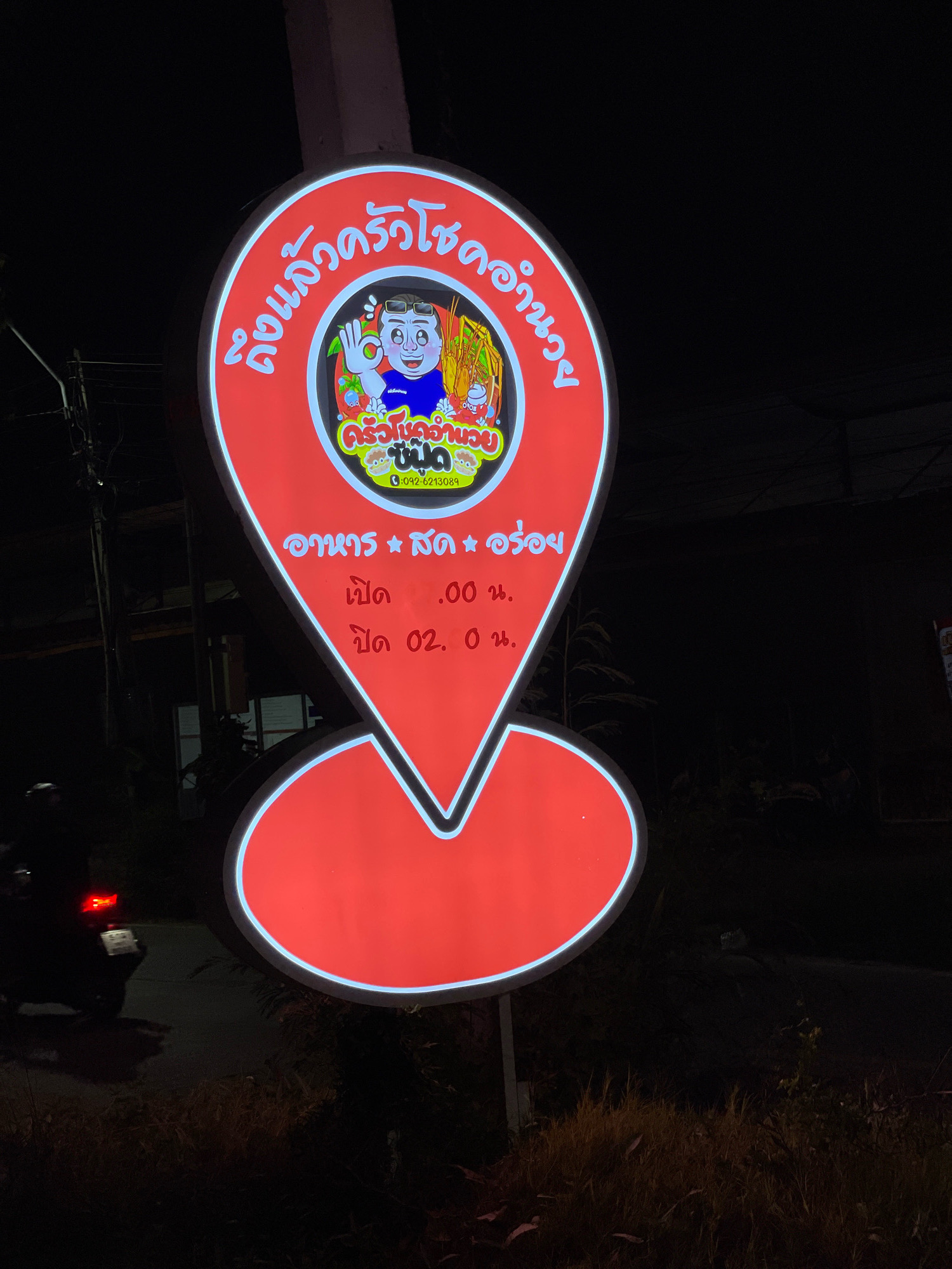 The image shows a brightly lit red and white sign in the shape of a map marker, advertising a restaurant named "ครัวคุณปู่" (Krua Khun Pu). The sign is illuminated at night. At the top of the sign, in a curved white font on a red background, are the words "ถึงแล้วครัวคุณปู่" (Reached Krua Khun Pu). In the center of the sign is a cartoon illustration of a man with sunglasses and a blue shirt, holding up an "ok" hand gesture, with a steaming bowl of noodles and shrimp in front of him. Below the illustration, in white font on a yellow and green background, is the restaurant name "ครัวคุณปู่" (Krua Khun Pu) and below that, the phone number "092-6213089". Further down, in white font on a red background, it lists "อาหาร * สด * อร่อย" (Food * Fresh * Delicious) and then the opening and closing times: "เปิด 11.00 น." (Open 11:00 AM) and "ปิด 02.00 น." (Close 2:00 AM). The sign is attached to a white pole and is located outdoors at night. In the background, a motorcycle is visible on the left side of the image, its taillight illuminated. The surrounding environment appears to be a roadside with some vegetation. This sign is located in Pattaya City, Thailand, as indicated by the context of the restaurant name and typical signage found in the area.