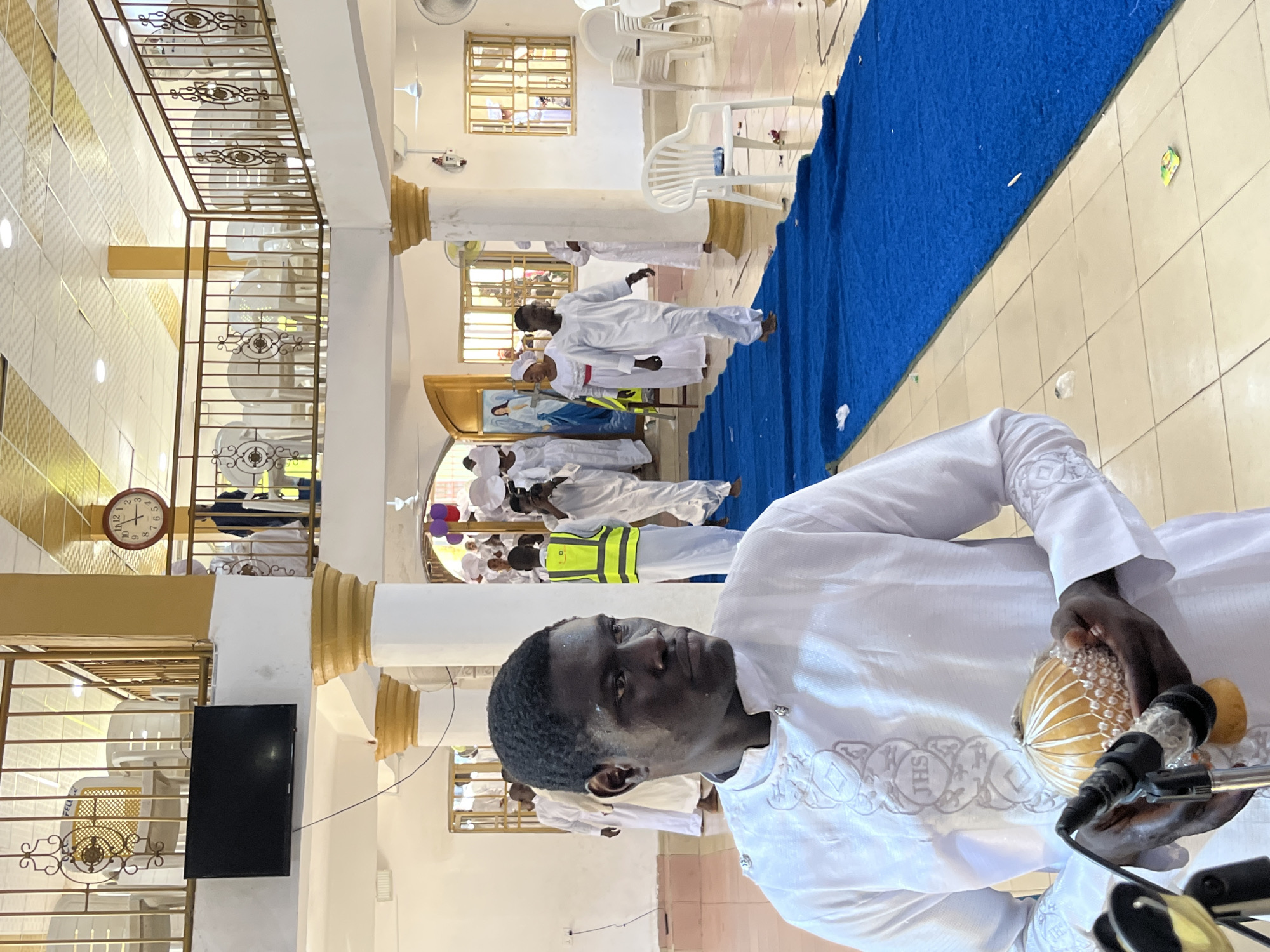 The scene depicts the interior of a large hall or church, in an unknown city and country. The multi-level structure features a main floor with light-colored square tiled flooring and a wide blue carpet runner extending from the mid-ground towards an arched doorway in the background. White, cylindrical pillars with gold-colored fluted capitals support the ceiling, which has patterned tiles, recessed lighting, and multiple white ceiling fans. A balcony with a brown metal railing, adorned with decorative scrollwork, overlooks the main floor.
Approximately 15-20 individuals are visible. In the lower-left foreground, a dark-skinned adult male stands, facing right with a slight turn of his head. He wears a long-sleeved white tunic with grey embroidered patterns, including the "JHS" monogram on the chest. He holds a light-colored, gourd-like instrument with white beads, resembling a shekere, in both hands, positioned near a microphone stand.
In the mid-ground and background, multiple dark-skinned adults are walking along the blue carpet and through an archway on the left. Most are dressed in white, flowing robes or tunics. One person in the mid-right wears a yellow high-visibility vest over white attire. Another individual further back is holding a camera. Through the archway on the left, a large painting or tapestry depicting a robed figure, predominantly in blue and white, is visible. One woman near this archway wears a white dress with a red trim.
On the upper level, several white plastic chairs are arranged behind the railing. A black flat-screen television is mounted on a white wall below the balcony railing on the left. A round wall clock with Roman numerals, displaying approximately 4:25, is mounted on a gold-colored wall section on the upper right. White plastic chairs are also scattered on the main floor to the right. Small pieces of debris, including wrappers and possibly flower petals, are present on the tiled floor and the blue carpet. Rectangular windows with light-colored grilles are visible in the background walls. Deflated or partially deflated balloons (purple and red) are near a pillar in the mid-right.