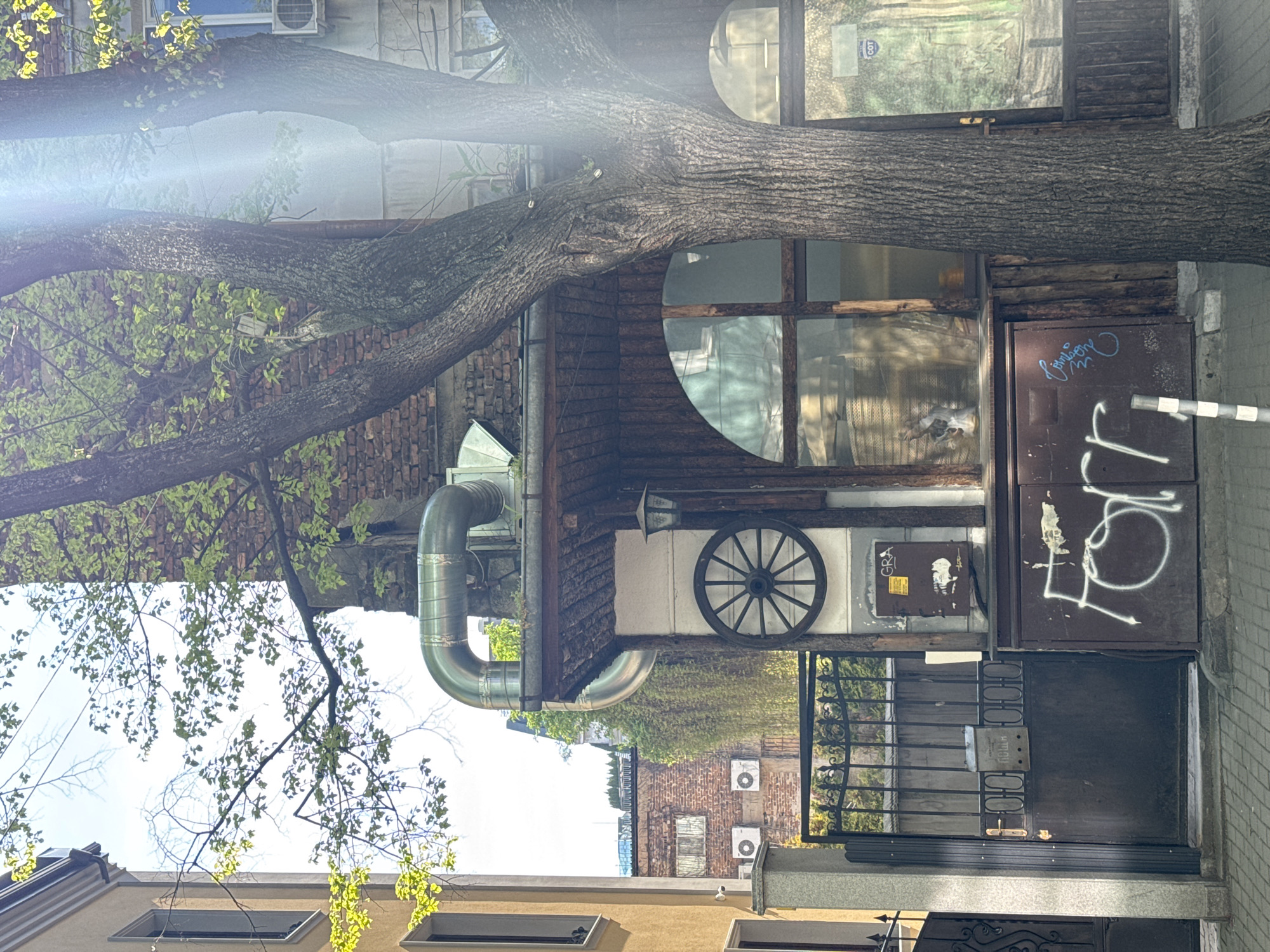 The image depicts a vibrant, sunlit street scene in an urban environment, likely a narrow passage or alleyway in Sofia, Bulgaria. A large, mature tree with rough bark and abundant fresh green leaves dominates the midground, its thick trunk stretching prominently across the frame.
On the right, a distinctive building features dark wooden siding and two striking circular windows. Above the lower circular window, a decorative wagon wheel is affixed to the wall, with a small light fixture above it. To the right of this wooden facade, a dark metal panel is covered with white graffiti, including a stylized "L" or "76" and a partially obscured cursive word. Below this, a black metal gate with an ornate design and a silver plate reading "МЕТАЛ" (Cyrillic for "METAL") provides entry.
On the left, a building constructed of reddish-brown brick is partially visible through the tree's foliage. Several rectangular windows are scattered across the buildings, some reflecting the bright sky. A large, shiny, corrugated metal pipe, possibly a ventilation or exhaust duct, curves prominently from the left side of the wooden-sided building. Multiple outdoor air conditioning units are mounted on the buildings in the background.
The scene is bathed in bright daylight, with a strong sun flare visible through the tree branches on the left, indicating a clear, sunny day, likely during spring or early summer due to the lush green foliage. Visible text includes "COT" on a window sticker and "GRA" on a brown electrical box, in addition to the graffiti and the "МЕТАЛ" sign. The overall aesthetic combines rustic elements with urban infrastructure.