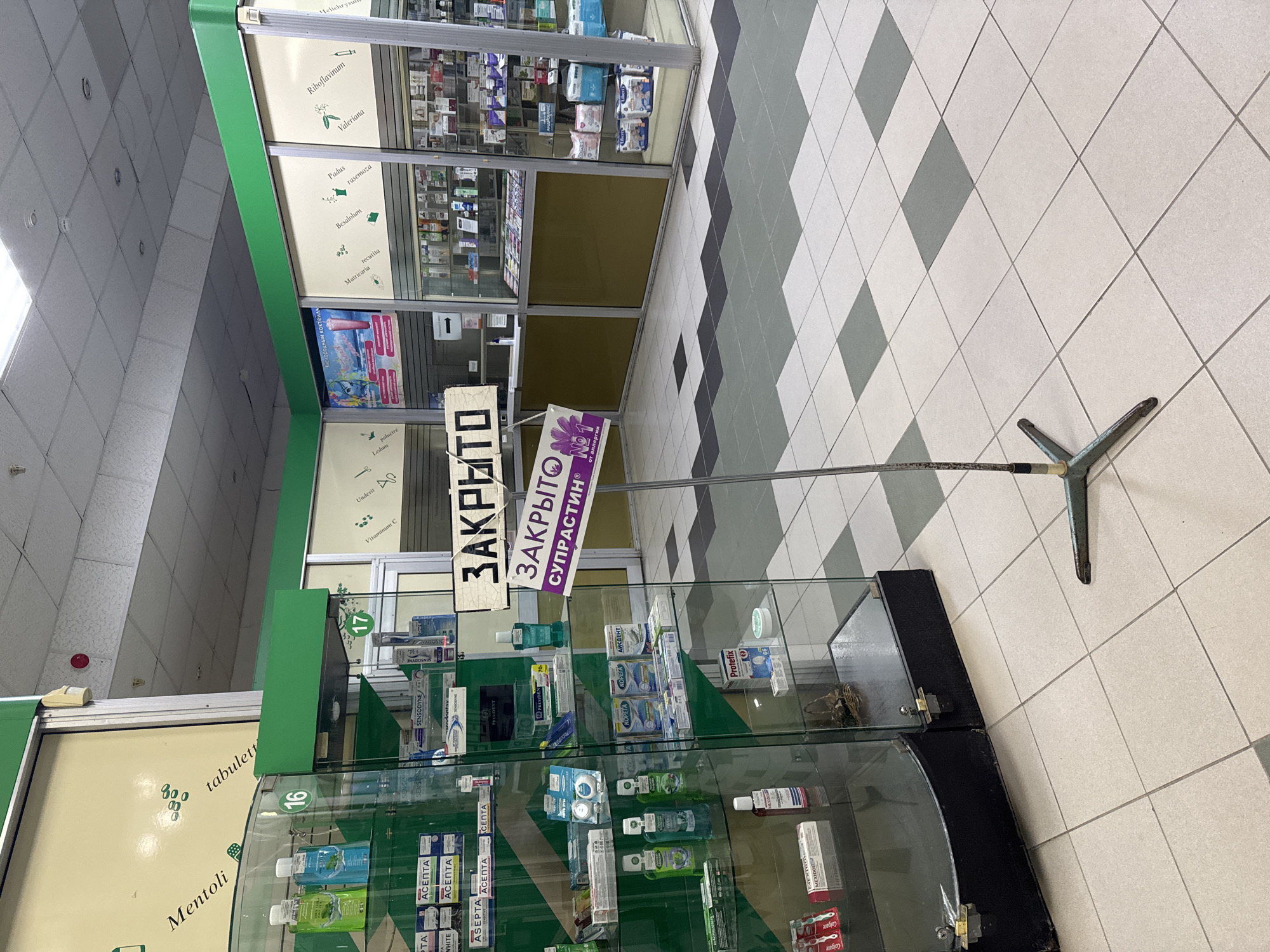 The image depicts the interior of a pharmacy. The scene includes shelves stocked with pharmaceutical products, displays with signage indicating that the checkout is closed ("ЗАКРЫТО"). The floor is tiled in a light and dark gray pattern. The overall setting suggests a retail environment focused on healthcare and personal care products.