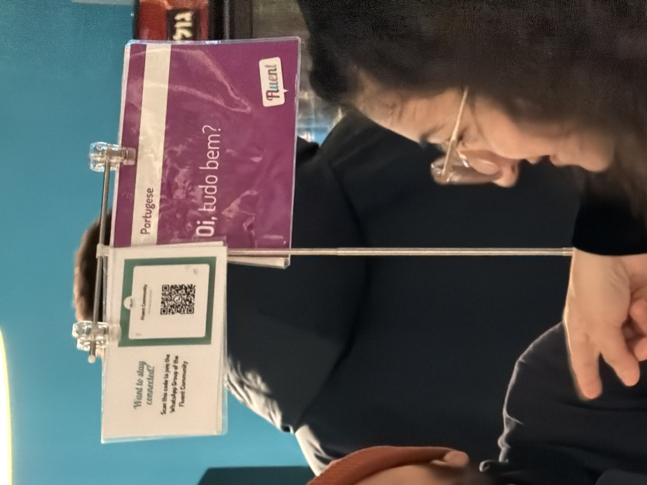 A language learning display features a card that says "Portuguese" and then "Oi, tudo bem?" which translates to "Hi, how are you?". The display also includes a QR code with text reading "Want to stay connected! Scan this code to join the WhatsApp Group of the Fluent Community". The word "Fluent" is visible on the purple card. In the background, people are interacting with the display.