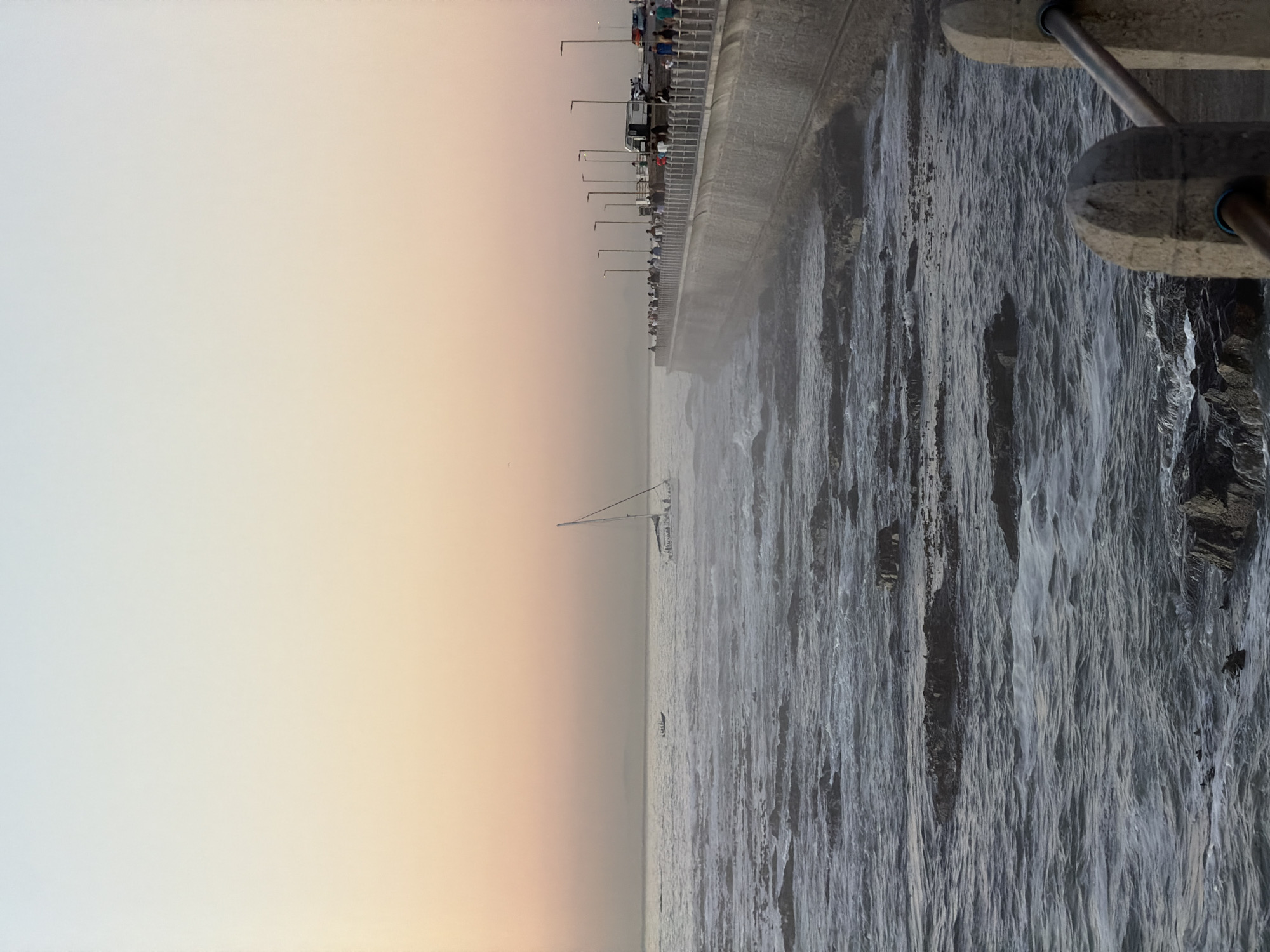 The image shows the coastline in Cape Town, South Africa. A pier is lined with people, and the sea is turbulent. A sailboat is visible on the horizon. A concrete barrier runs along the edge of the pier. The sky is a pale gradient of white and peach.