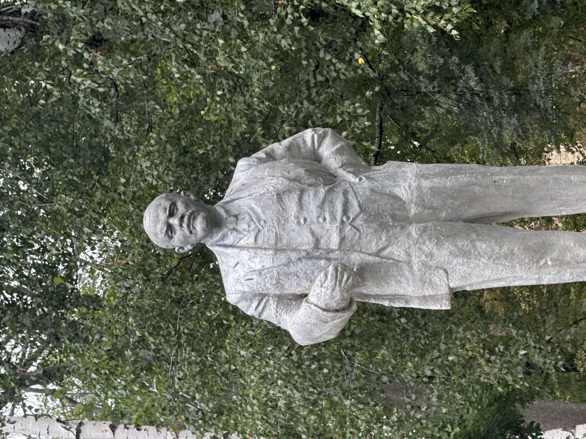 A full-body, slightly low-angle, front-facing outdoor shot features a weathered, light-gray concrete statue of Vladimir Lenin standing in front of a dense backdrop of green foliage. The statue depicts Lenin in a suit and waistcoat, with his right hand resting on his hip and his left arm bent with his hand near his chest. His head is slightly tilted forward, and he has a mustache. The statue is textured, showing the rough application of the concrete. The background consists of numerous leaves and branches of trees, with some lighter patches of sky visible through the canopy. The overall setting appears to be a park or a public square in Seleznivka, Ukraine, suggested by the presence of a statue of historical significance in such a location.