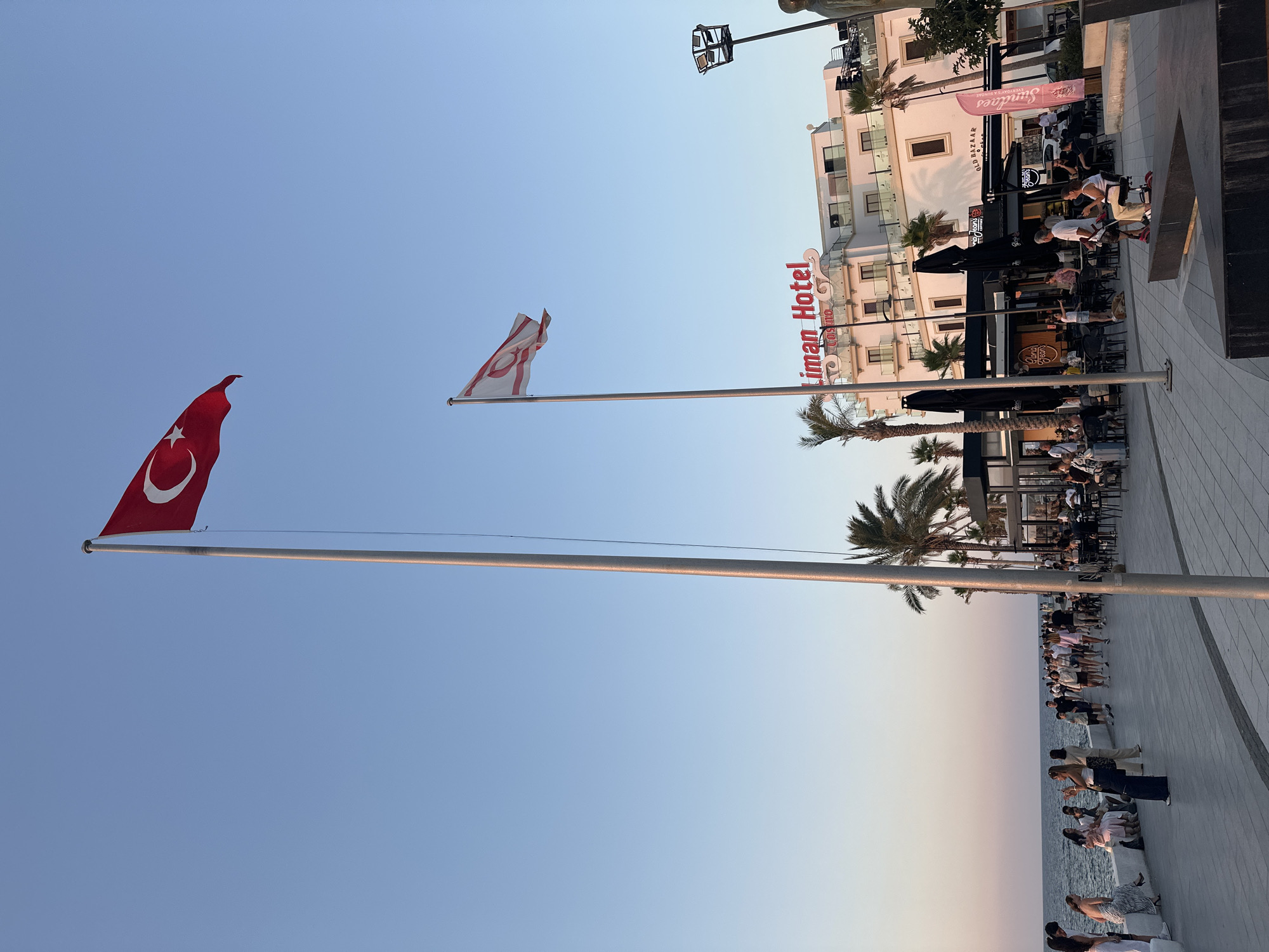 This is a photograph taken at sunset in the city of Kyrenia, Northern Cyprus. Two flags are prominently displayed on tall flagpoles: the Turkish flag, a red flag with a white crescent moon and star, and the flag of the Turkish Republic of Northern Cyprus, a white flag with two red horizontal stripes and a red crescent moon and star. In the background, a white building with "Liman Hotel" and "Casino" written in red lettering is visible. The building has multiple floors with balconies and windows. In front of the hotel, there are several outdoor seating areas with tables and chairs, suggesting a restaurant or cafe. Palm trees are scattered throughout the scene. A paved walkway runs along the coastline, with a body of water, presumably the sea, to the left. Numerous people are walking along the promenade and seated at the outdoor tables. The sky is a clear, pale blue, transitioning to a soft orange and pink hue near the horizon, indicating sunset. The overall scene depicts a lively waterfront area with a clear indication of the location through the flags and the hotel signage.