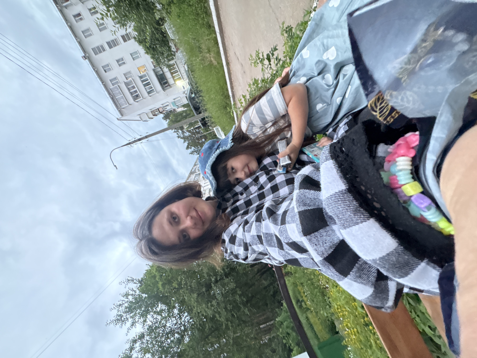 A medium shot captures two females, likely a mother and daughter, sitting on a bench outdoors in an urban setting. The woman in the foreground, on the left, has dark brown hair and is wearing a black and white checkered shirt. She is holding a young girl, who is on her right. The girl has long dark hair and is wearing a blue bucket hat with red embroidered stars and a striped t-shirt. She is holding a small box. The background shows a white apartment building with many windows, trees, and a street. The sky is overcast and cloudy. The focus is on the two individuals, with the environment serving as a backdrop.