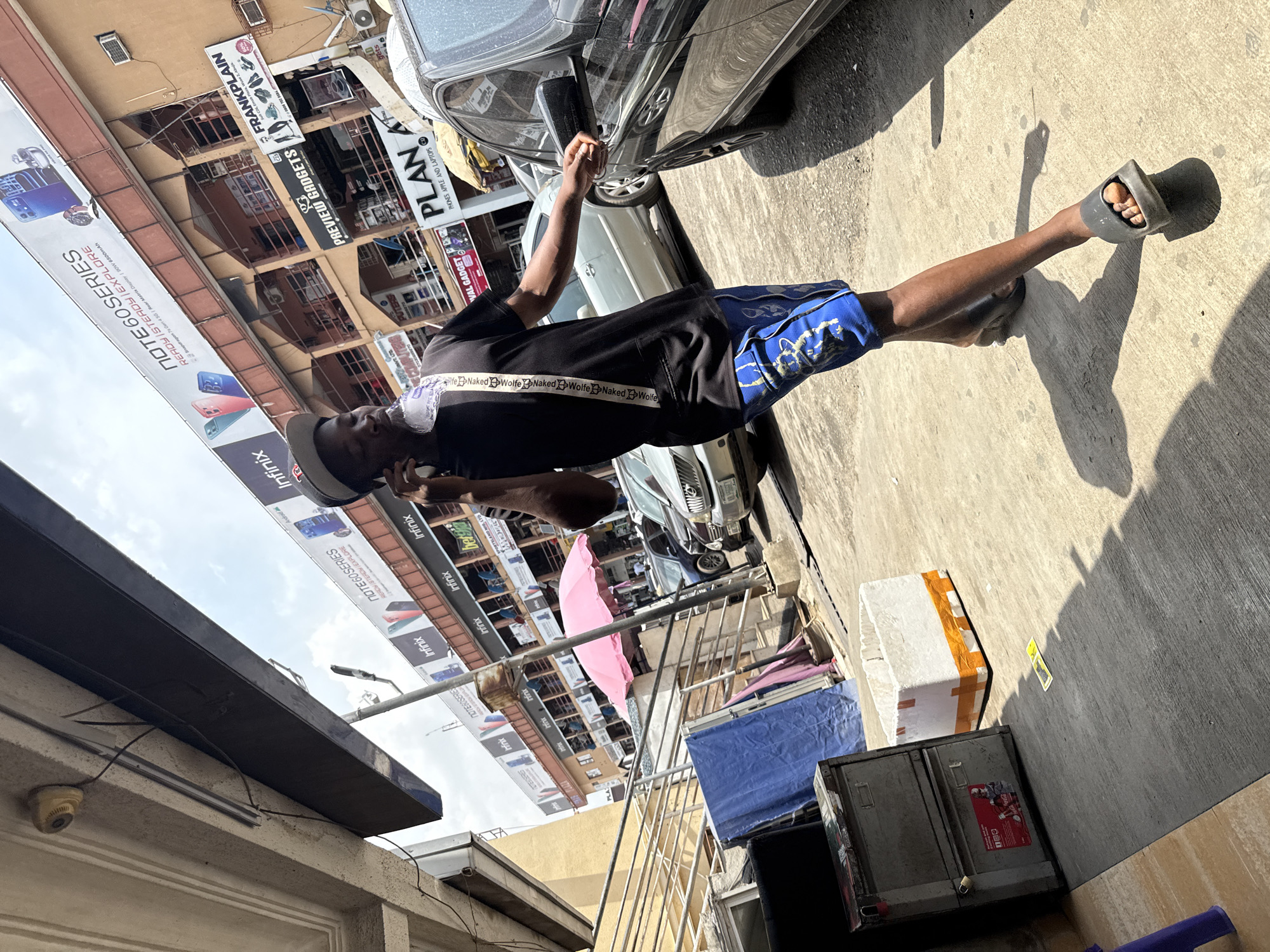 This media file captures a bright, sunny urban street scene in Abuja, Nigeria. A young man with dark skin stands in the middle, facing left. He wears a black t-shirt with a white band featuring "Naked Wolfe" text, blue and black patterned shorts, a grey cap, and grey slides/sandals. He is holding a phone to his right ear, seemingly in conversation, while his left hand holds a black object, possibly a car mirror cover.
The background features multi-story commercial buildings and parked cars, including a dark grey sedan. Numerous advertising banners are prominently displayed, primarily for "Infinix NOTE60SERIES" phones, detailing features like "READY STEADY EXPLORE," "Snapdragon 7s Gen 4SG," "Rear Matrix Display," and "90W 6500mAh." Other visible business names include "FRANKPLAIN," "PREVIEW GADGETS," "WAL GADGET," and "PLAN PHONES APPLE AND LAPTOPS." The text "Android" and "SPORTSLAND NIS LTD" are also visible on banners.
Strong shadows on the concrete ground indicate bright midday or afternoon sunlight. Objects in the scene include a metal cabinet, a white box with orange tape, a pink umbrella, a blue tarp, railings, and a surveillance camera mounted on a building overhang. The sky is partly cloudy.